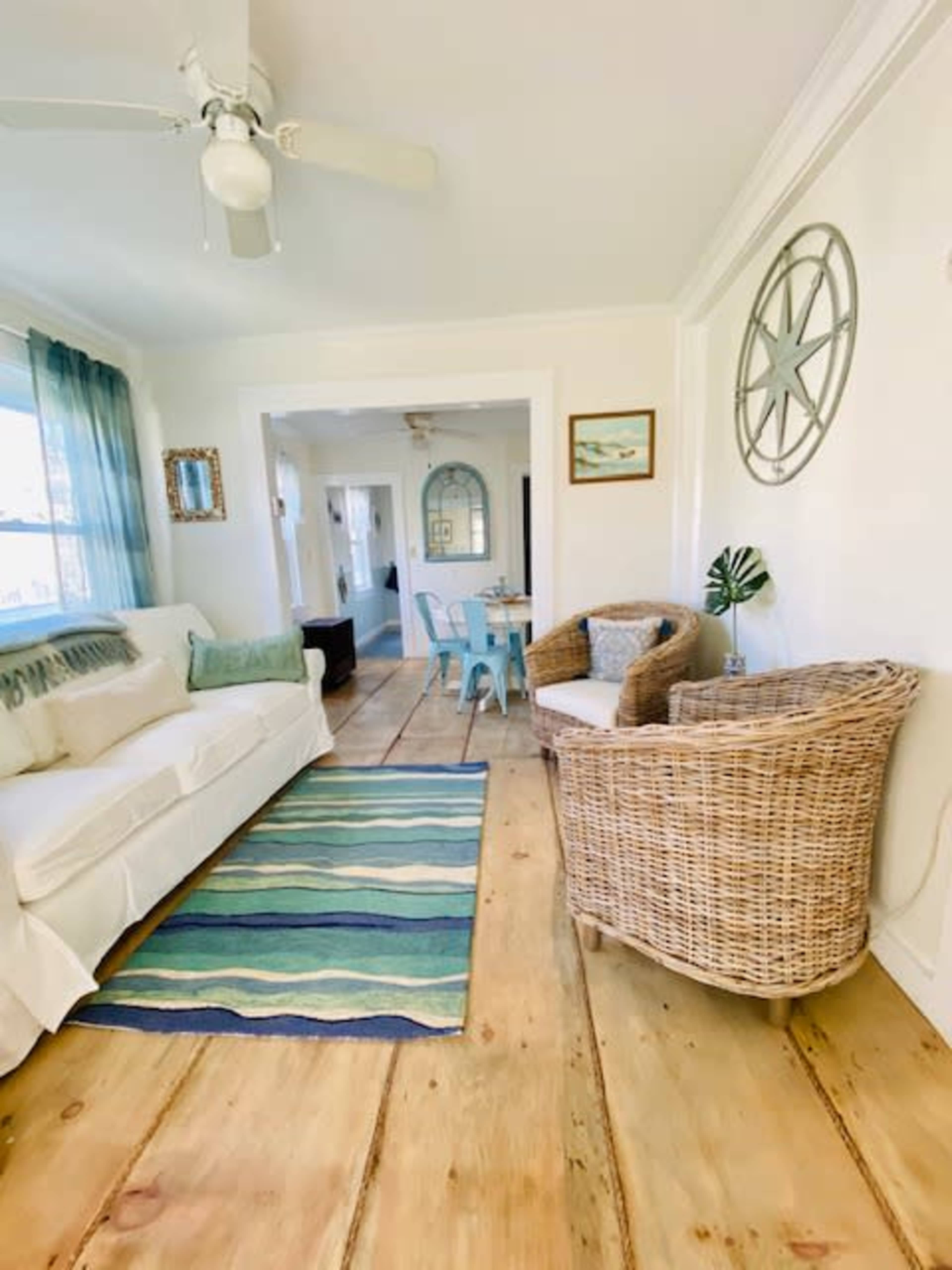 A light-filled living room with a white sofa, a striped rug, and wicker furniture, leading to a dining area in the background.