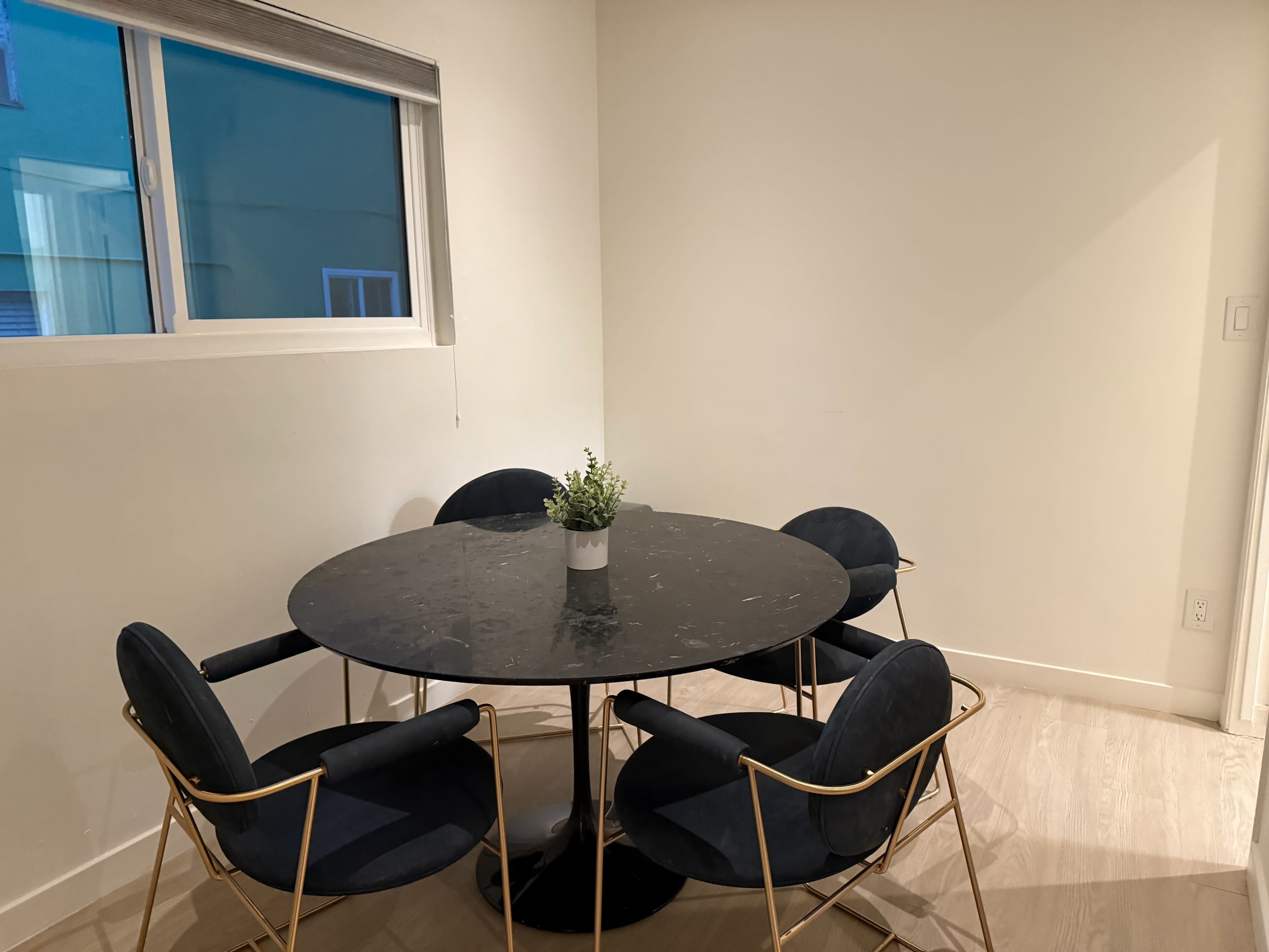 A round black marble table with gold metal legs is surrounded by four navy blue upholstered chairs in a well-lit corner of a room.