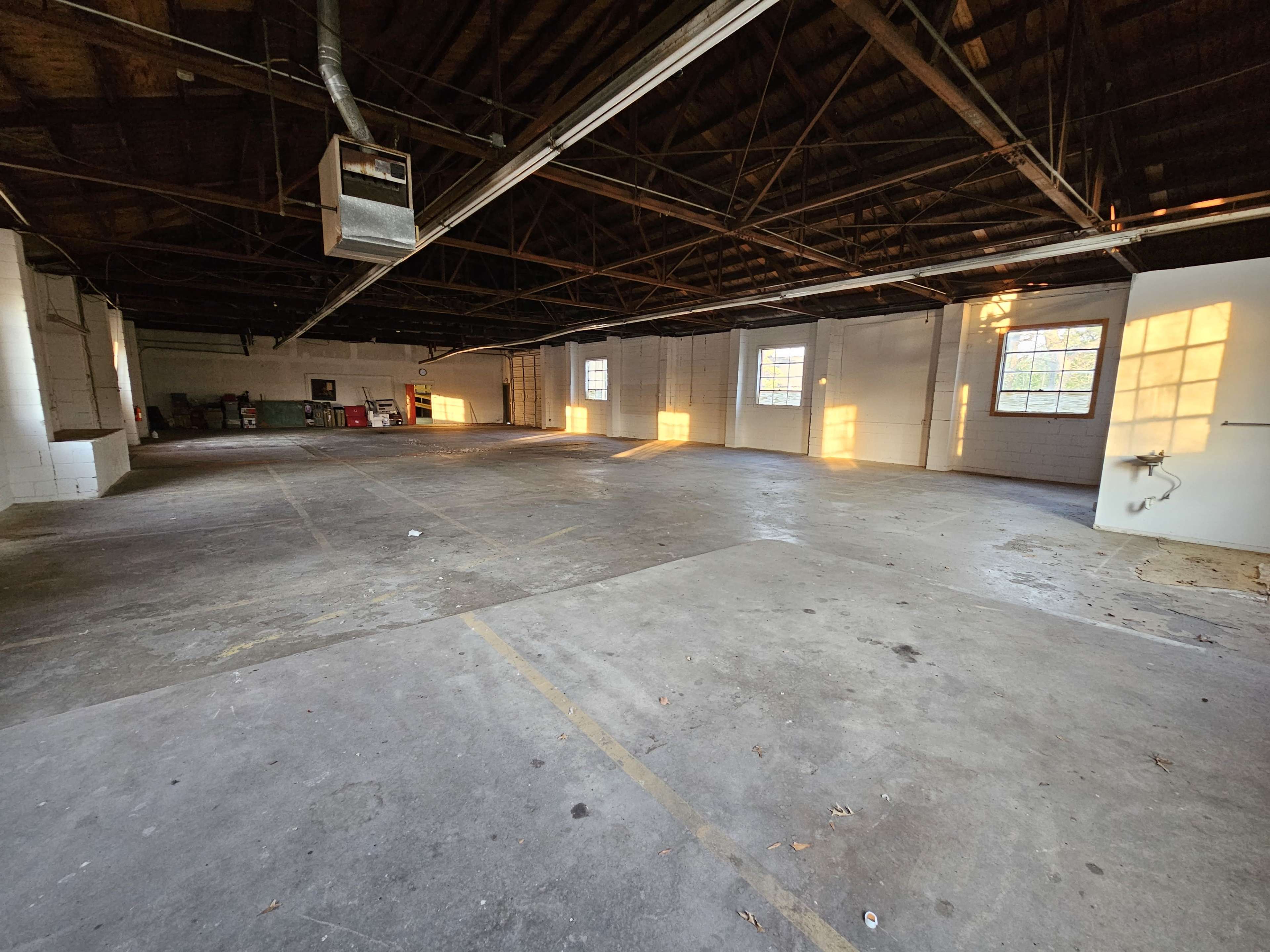An empty warehouse with exposed beams, concrete flooring, and sunlight casting shadows through the windows.