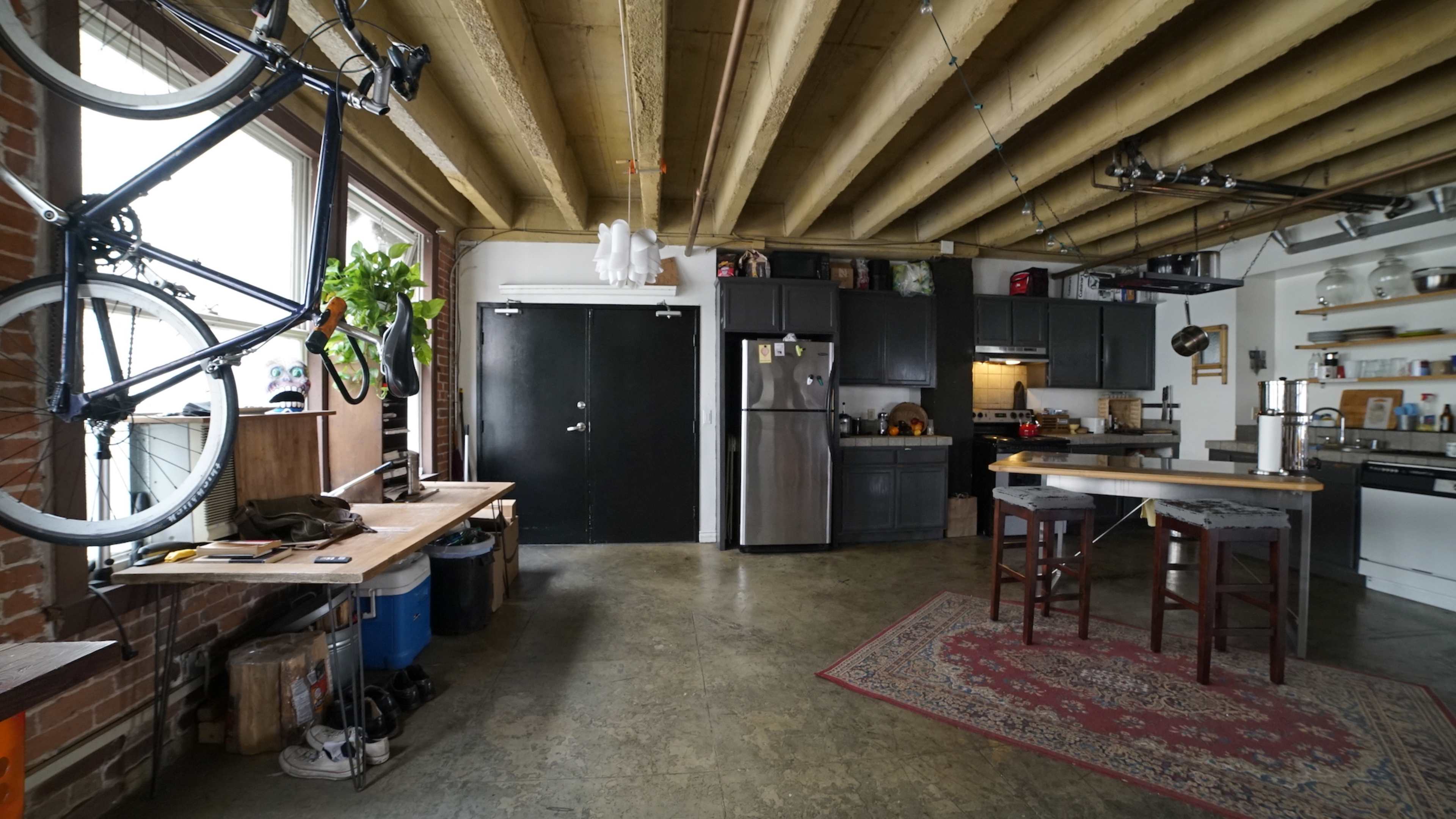 A spacious industrial-style kitchen and living area features exposed wooden beams, a bicycle mounted on the wall, and a kitchen equipped with stainless steel appliances.