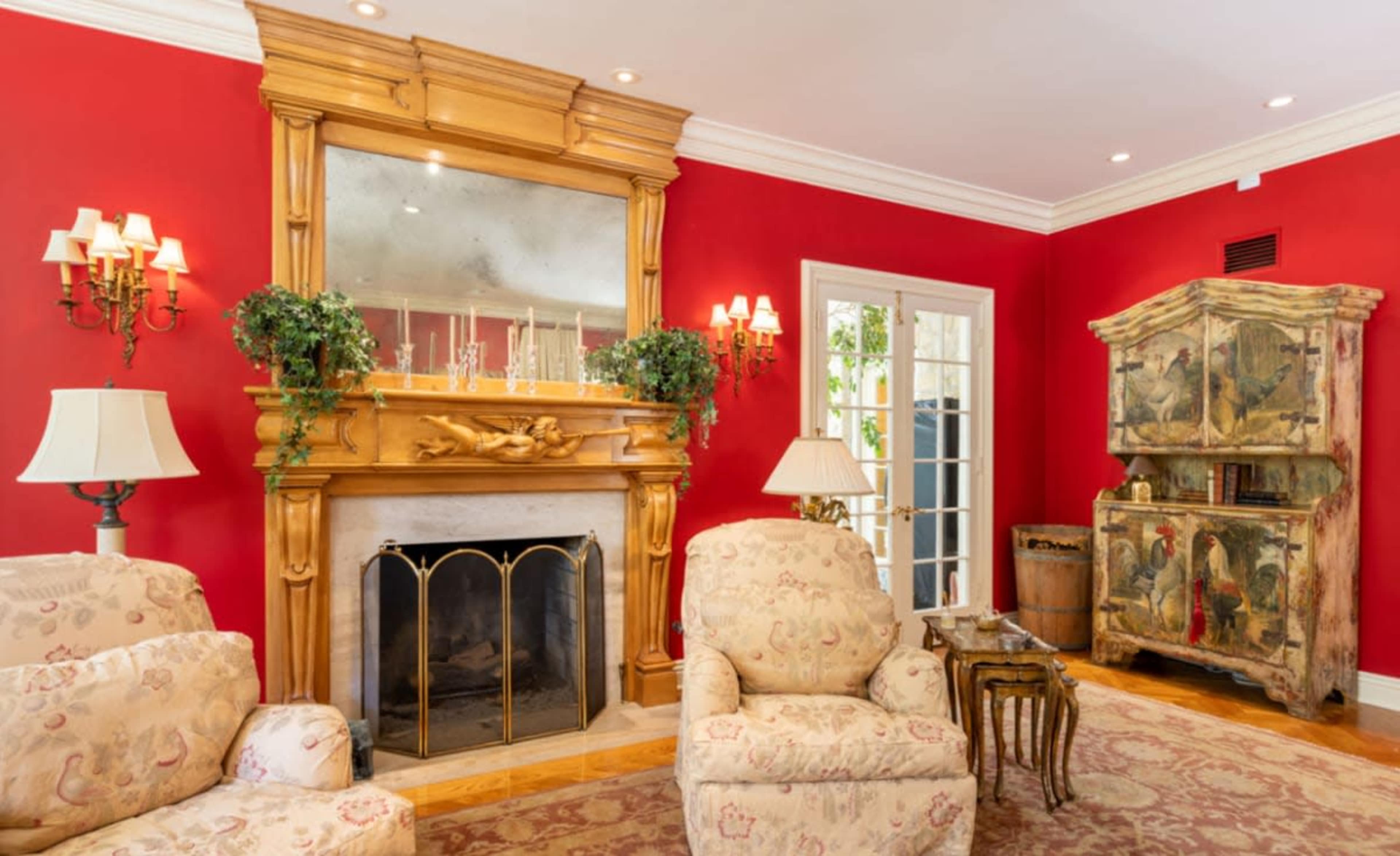 A living room with red walls, a wooden fireplace with a large mirror above it, and vintage furniture, including upholstered chairs and a decorative cabinet.
