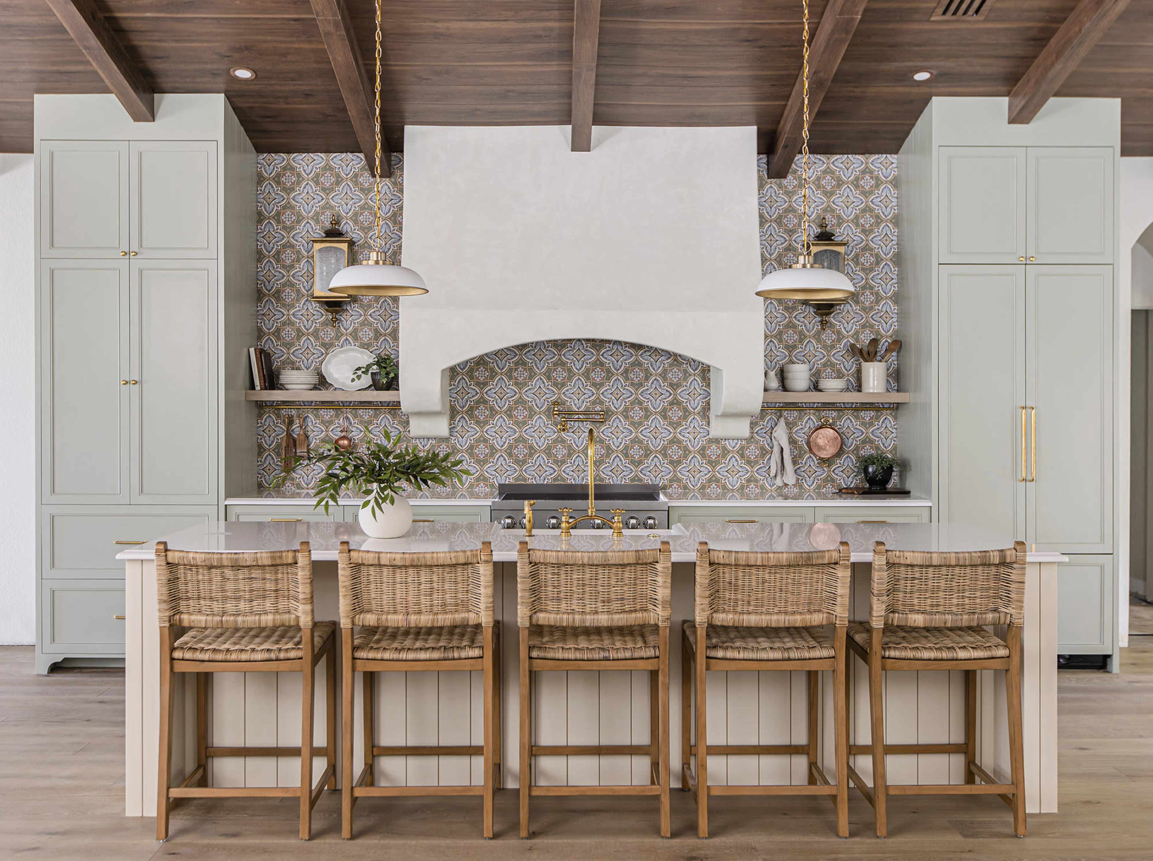 The kitchen features a large central island with woven bar stools, surrounded by light green cabinetry and intricate tile backsplash, complemented by a decorative range hood.