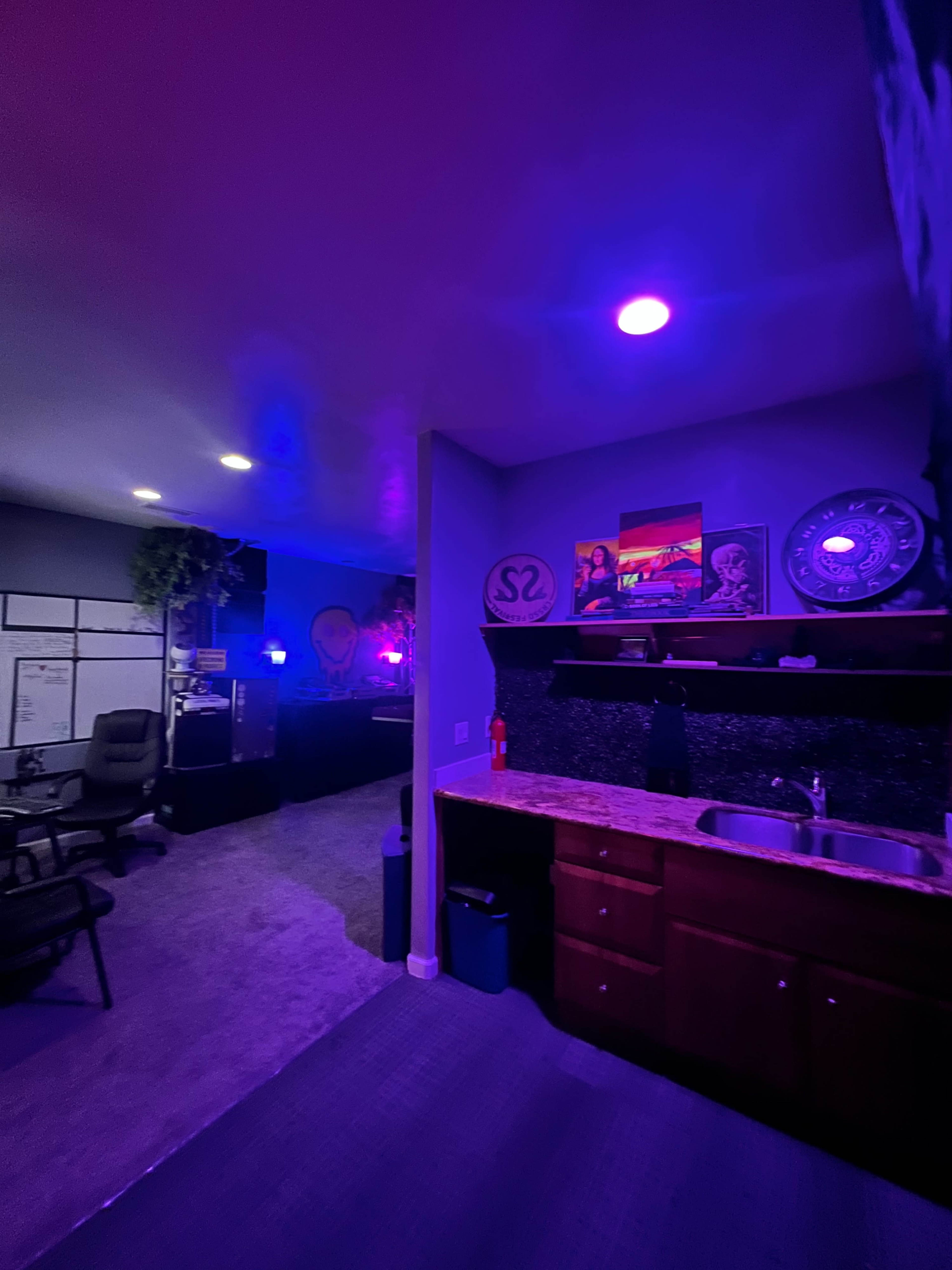 The image shows a modern workspace with a small kitchen area, illuminated by purple and blue lights, and a black office chair near a counter with a sink.