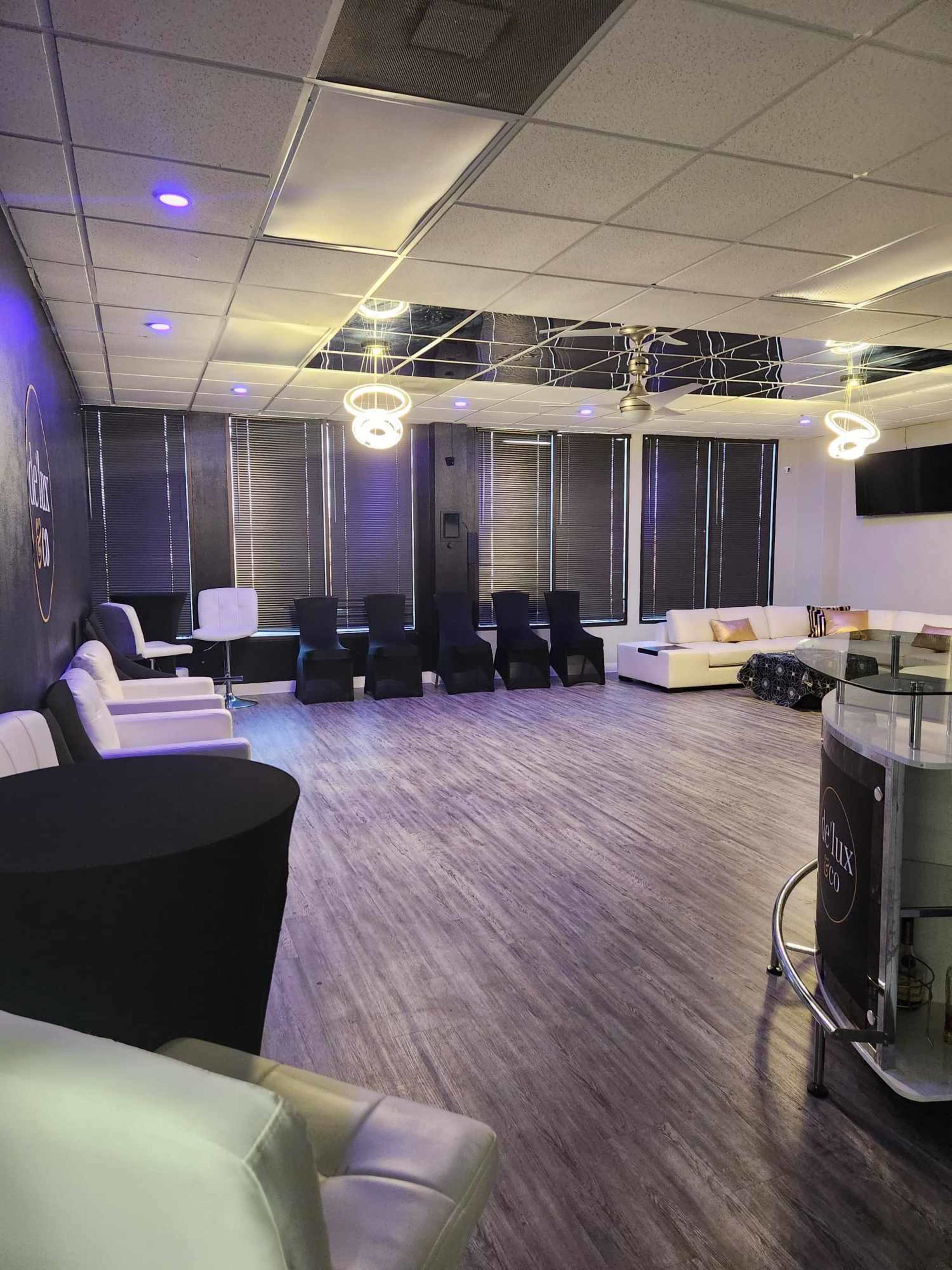 A spacious room with modern seating arrangements, featuring white couches and black chairs, illuminated by hanging lights and equipped with large windows covered by blinds.