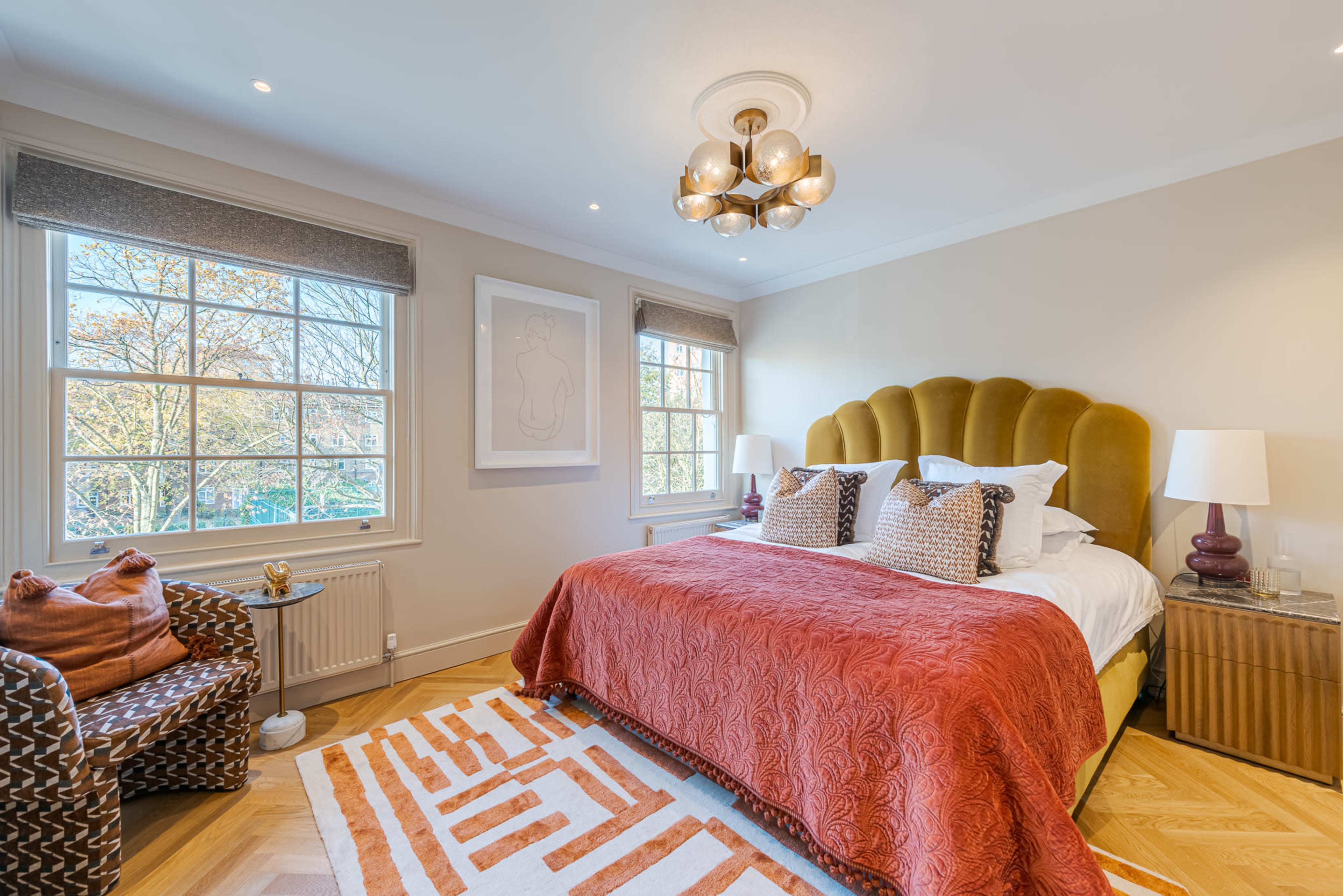 The image shows a modern bedroom featuring a large bed with a tufted headboard, accented by orange and neutral tones, along with two windows and a cozy seating area.
