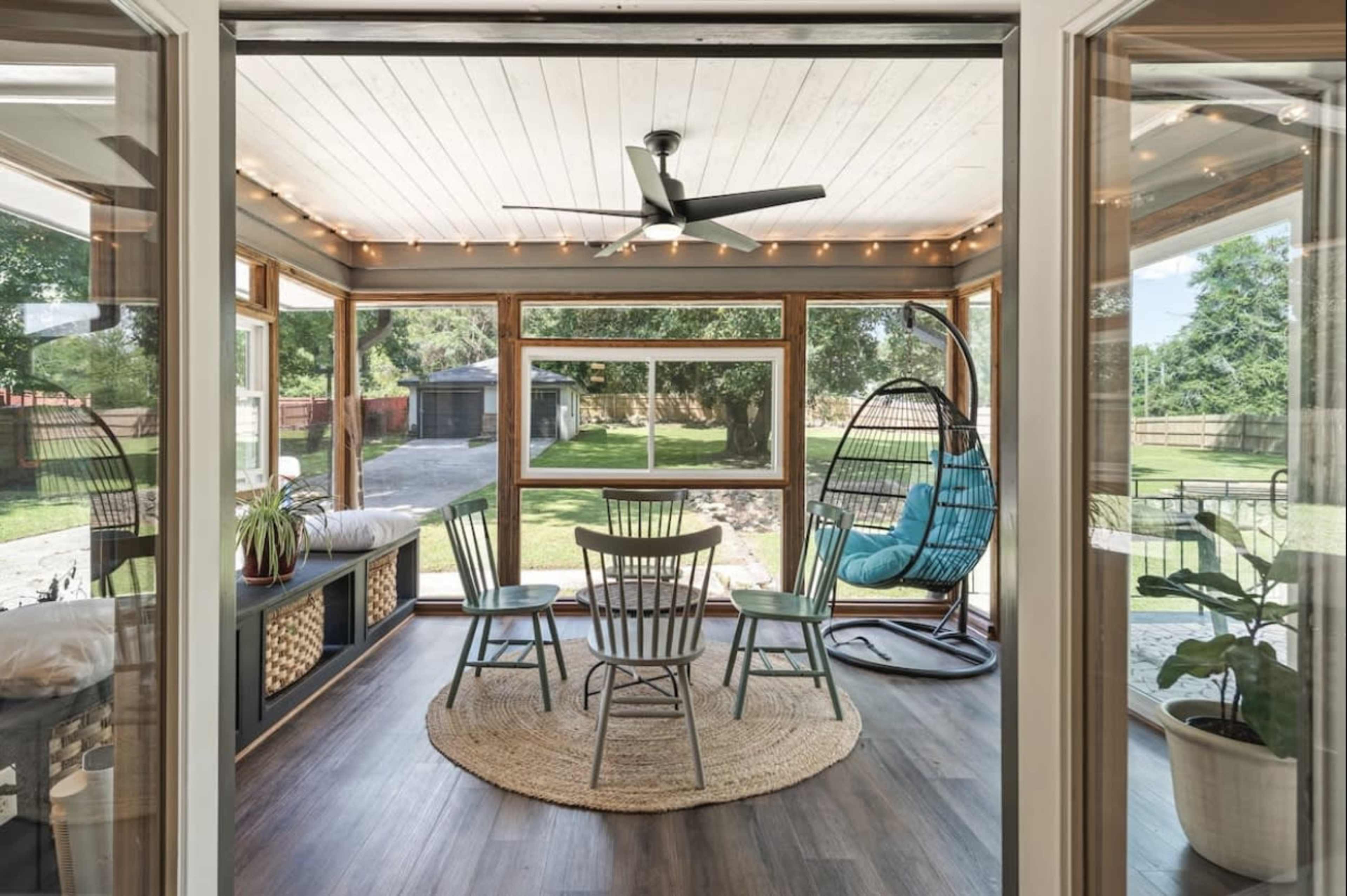 A bright sunroom featuring a circular rug, four wooden chairs arranged around a table, and a hanging chair beside large windows overlooking a garden.