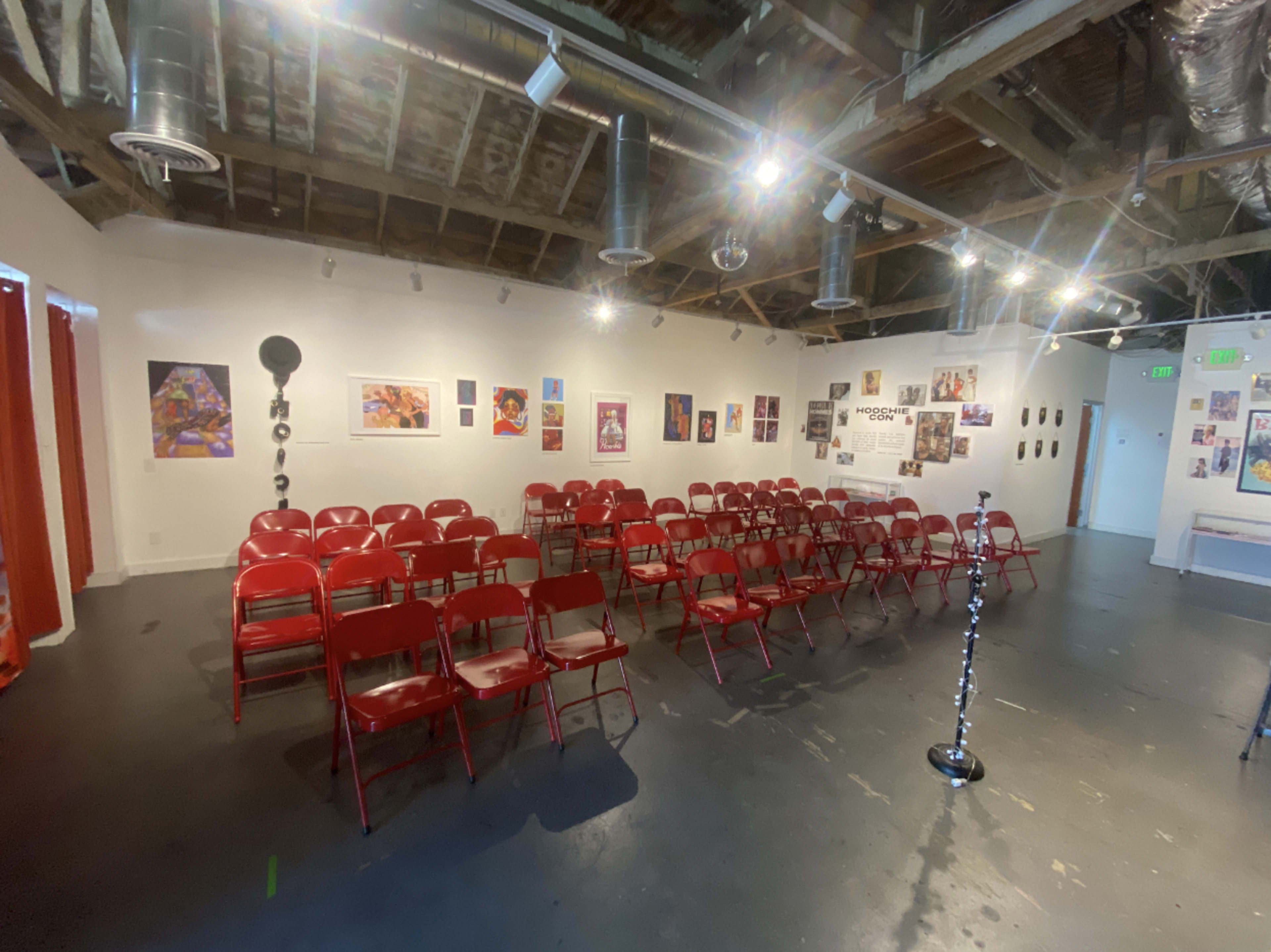 A vacant art gallery or performance space with rows of red chairs arranged in front of a series of framed artworks on the walls.