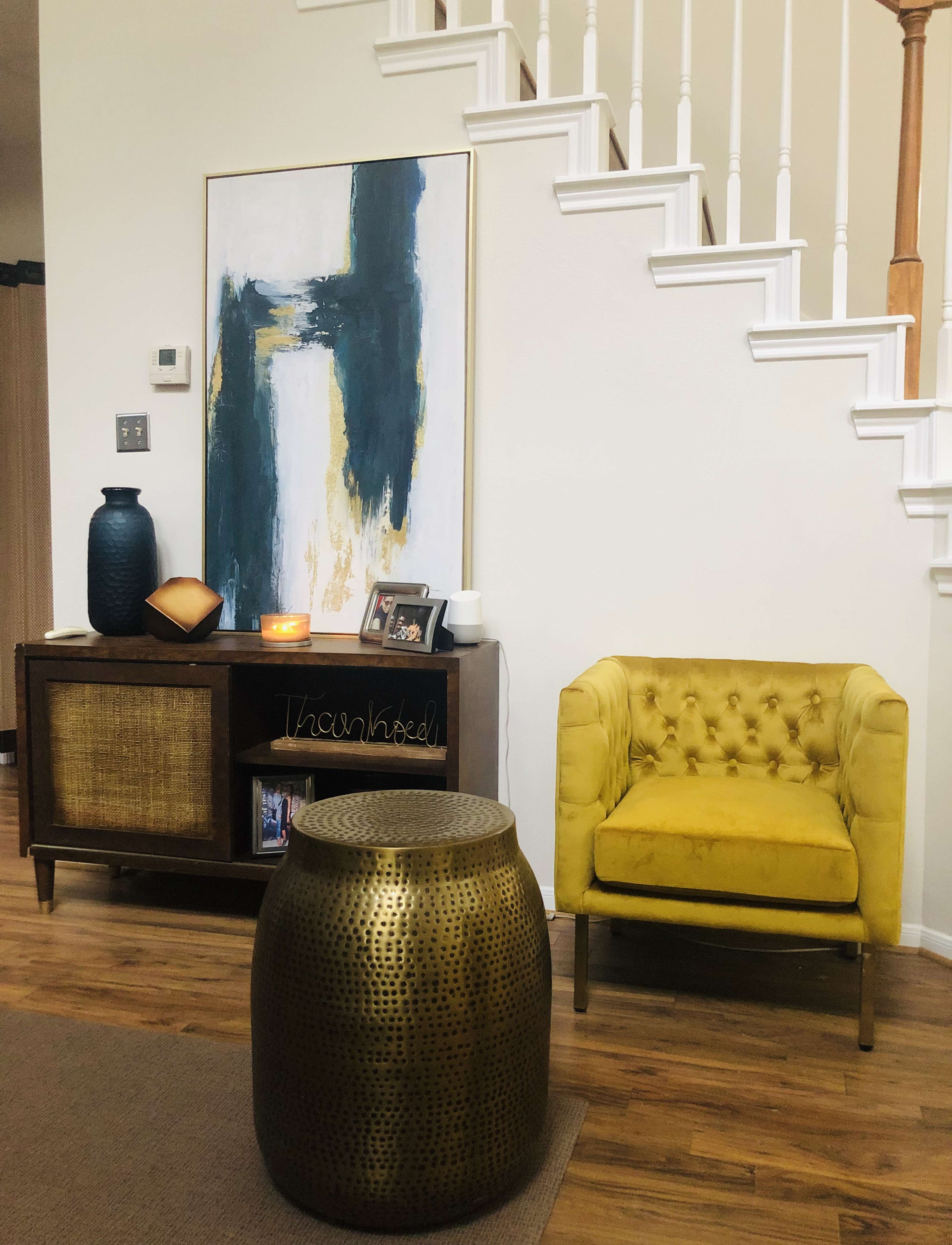 The scene features a yellow upholstered chair next to a wooden cabinet, a round brass accent table, and artwork on the wall, all set in a well-lit living room with a staircase in the background.