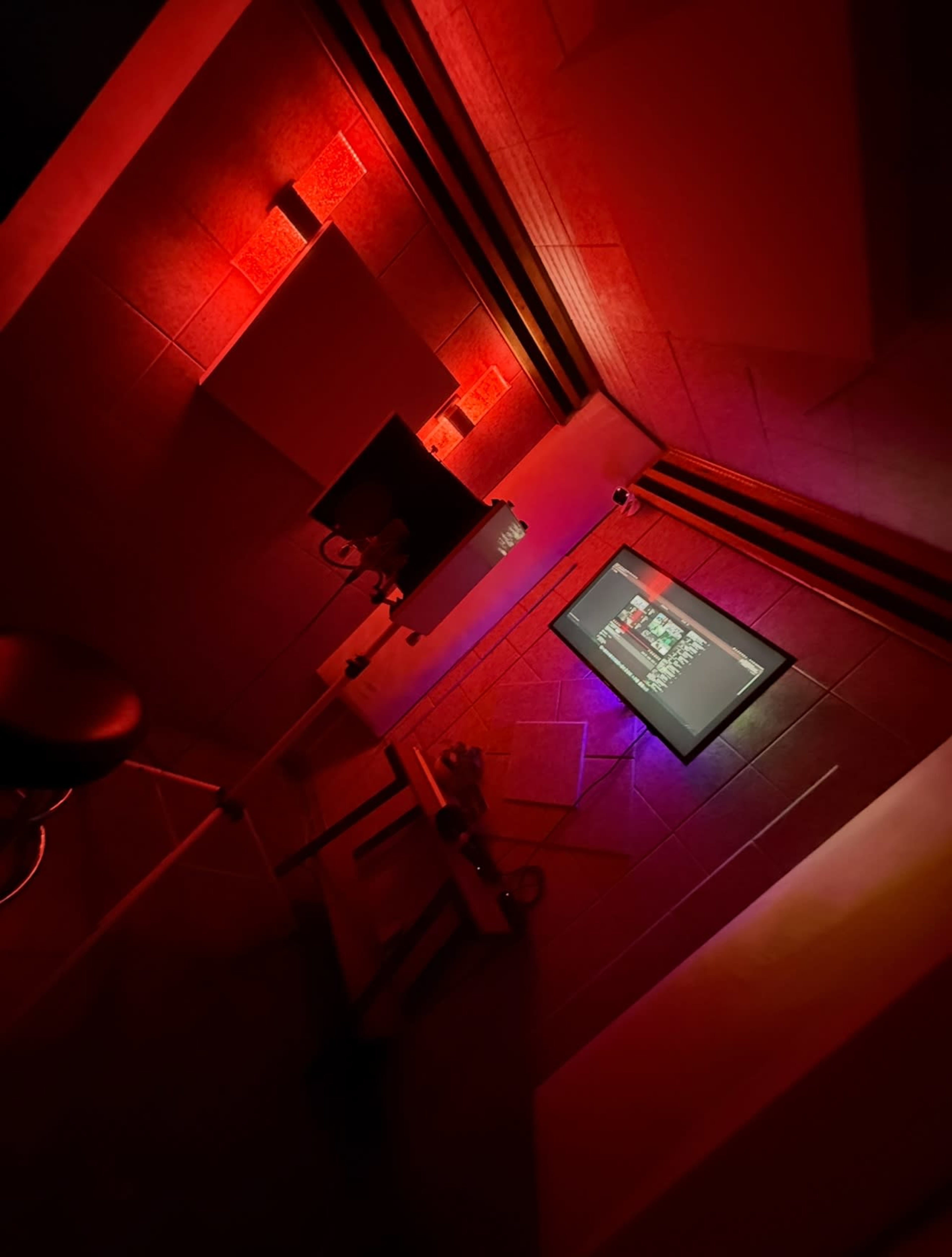 The image shows a dimly lit recording studio with red lighting, featuring a microphone, a monitor displaying audio settings, and equipment arranged in a corner.