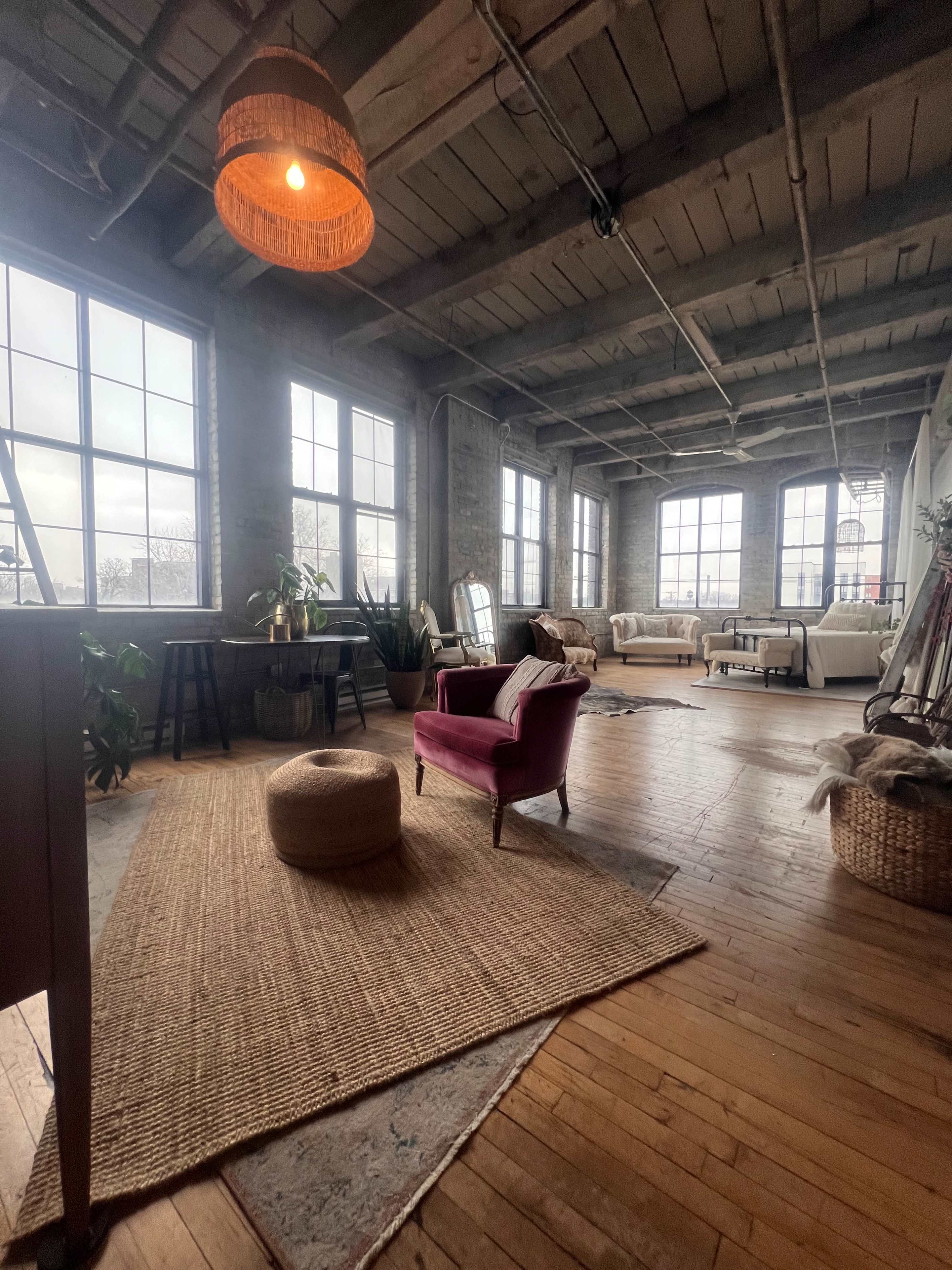 A spacious, industrial-style living area with large windows, a pink armchair, a jute rug, and various plants.