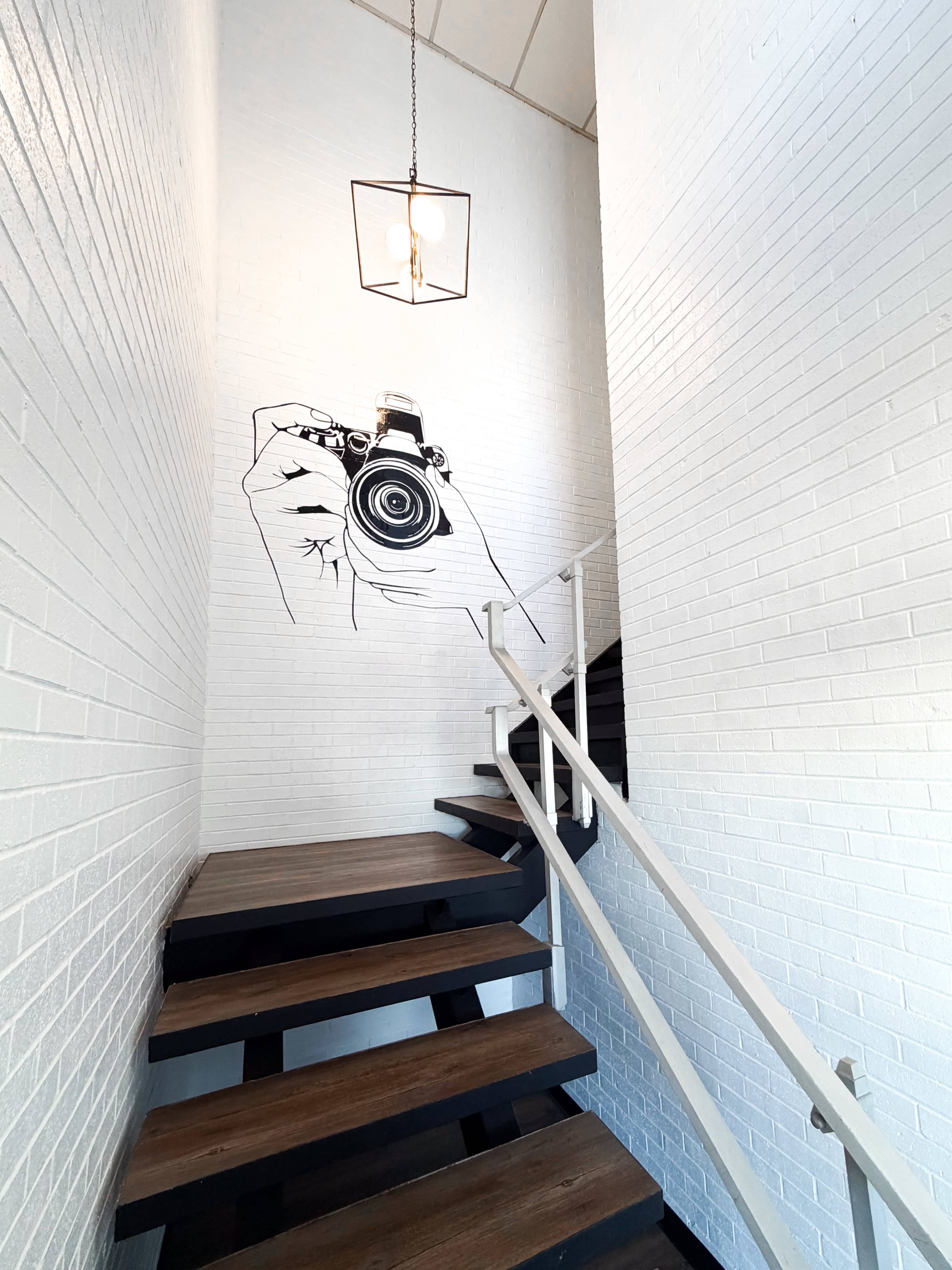 A hand-drawn mural of a camera adorns the white brick wall beside a wooden staircase leading upward.