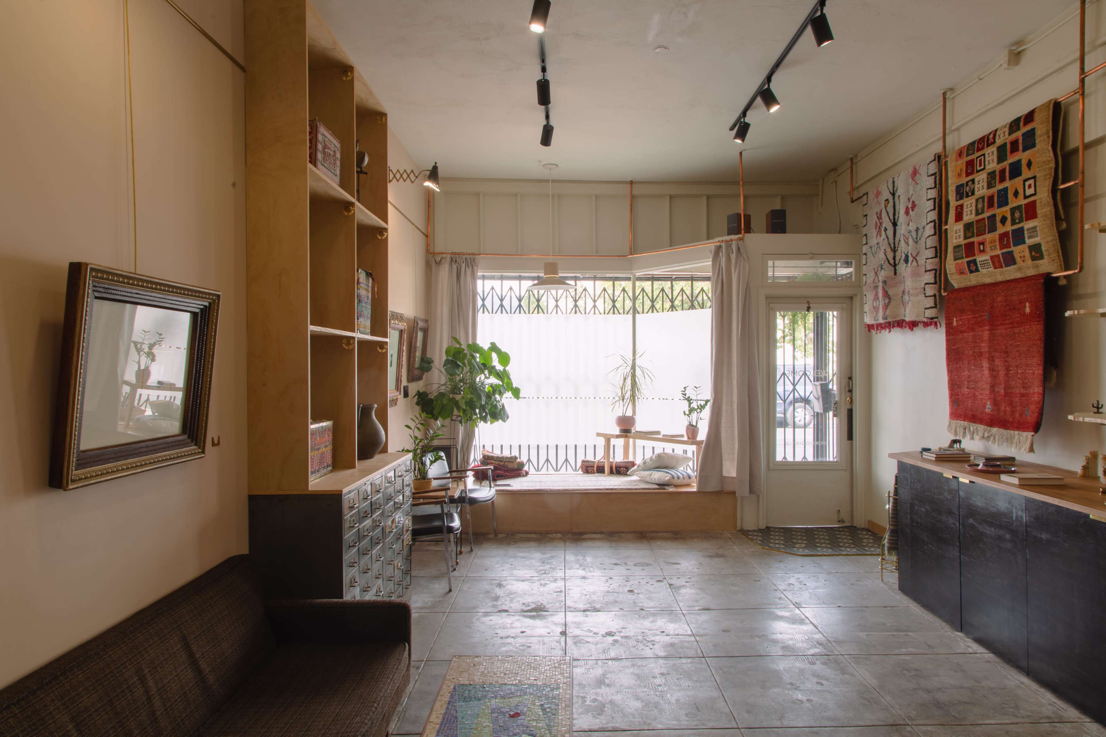 A cozy interior of a room with a sofa, bookshelves, and a large window, featuring plants and light-colored decor.