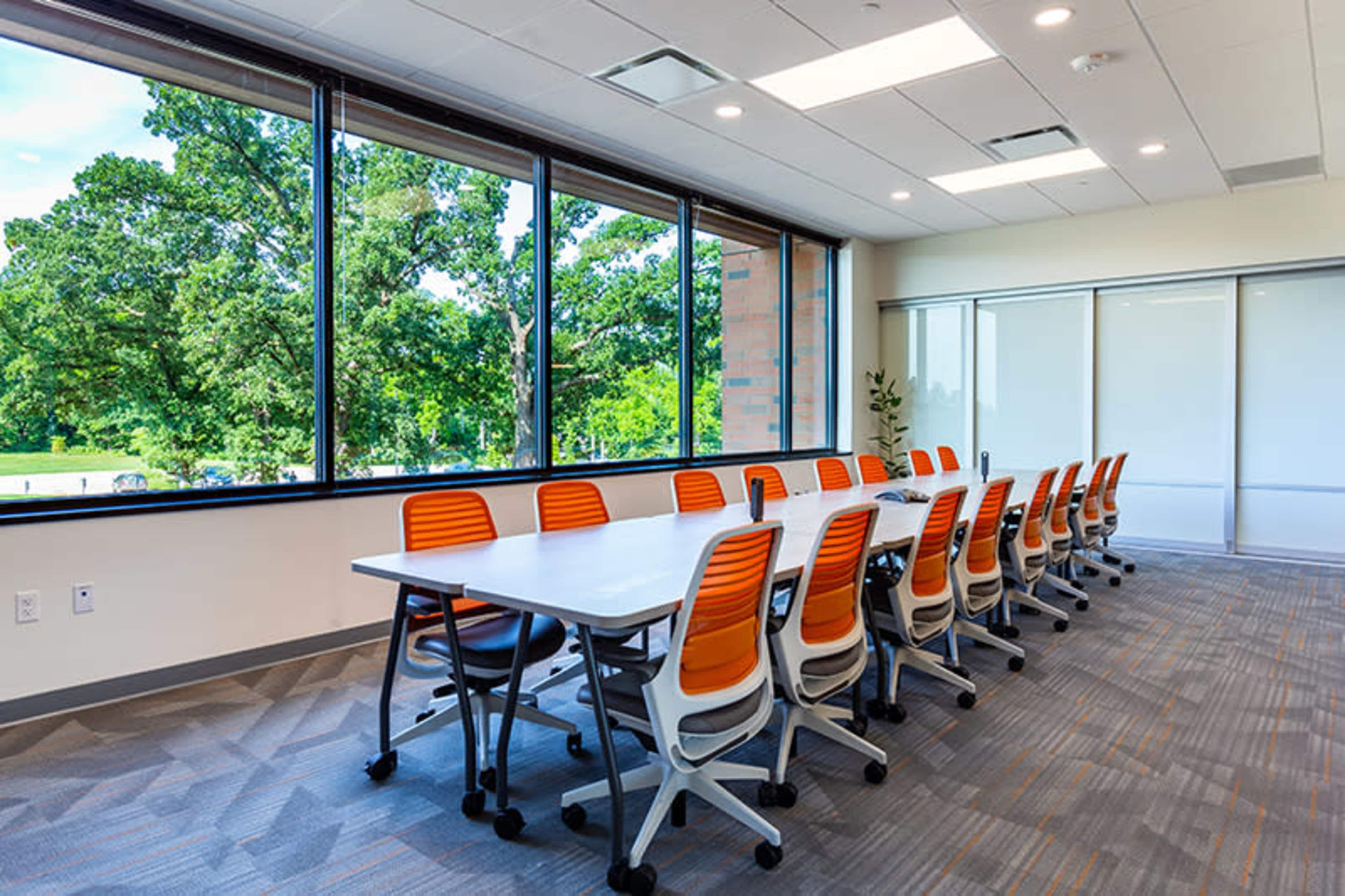 A conference room with a long table surrounded by orange and white chairs, featuring large windows that provide natural light and a view of greenery outside.