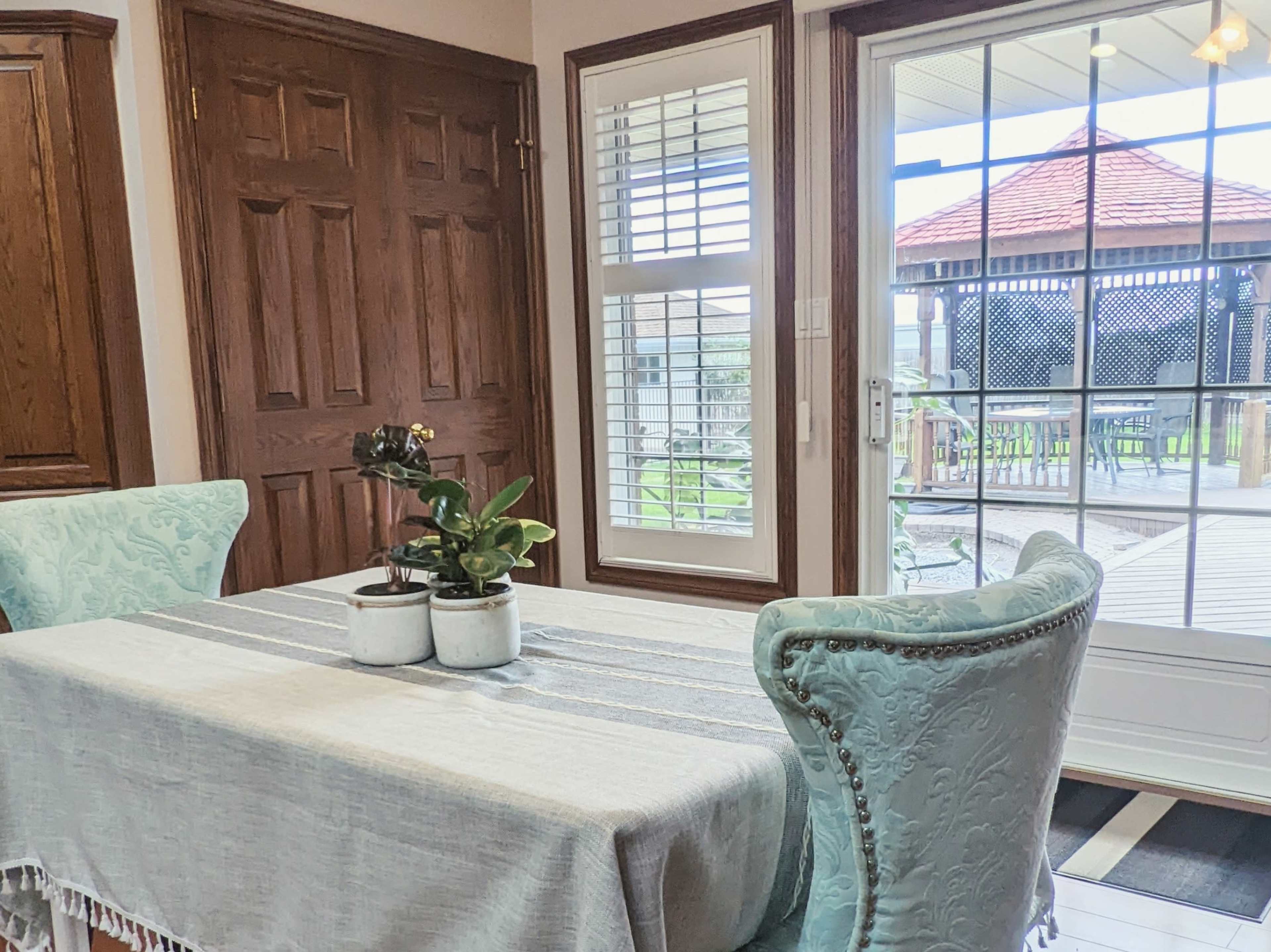 A dining table with a light gray tablecloth and two upholstered chairs is positioned near a glass door overlooking a deck with a gazebo.