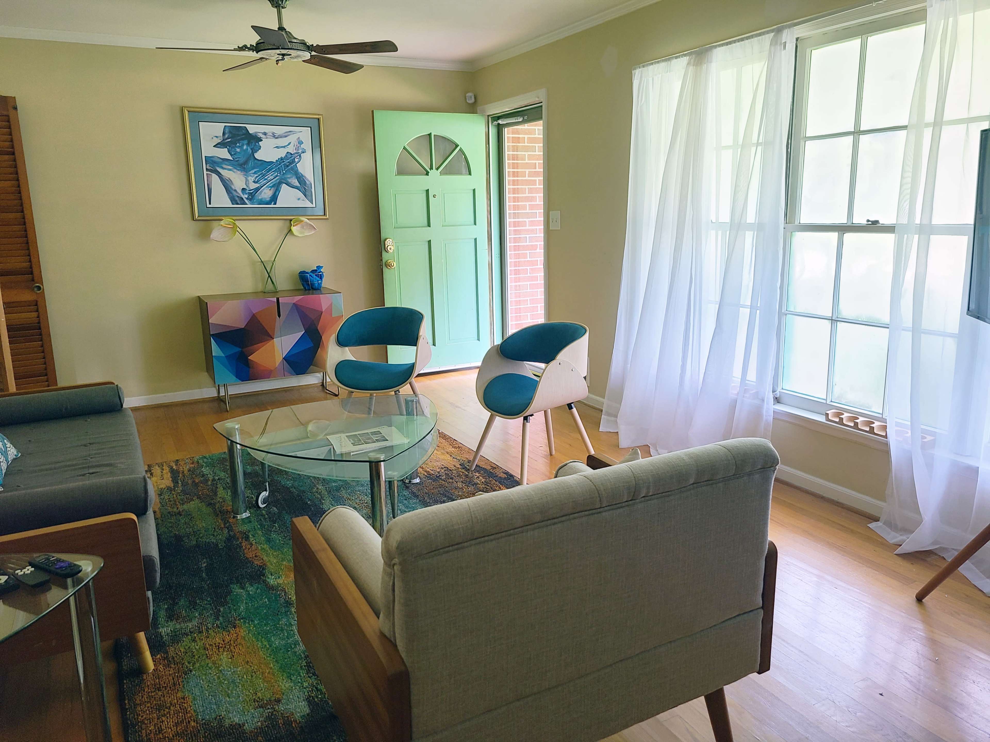 The living room features a coffee table, two modern chairs, a colorful artwork on the wall, and a large window with sheer curtains letting in natural light.