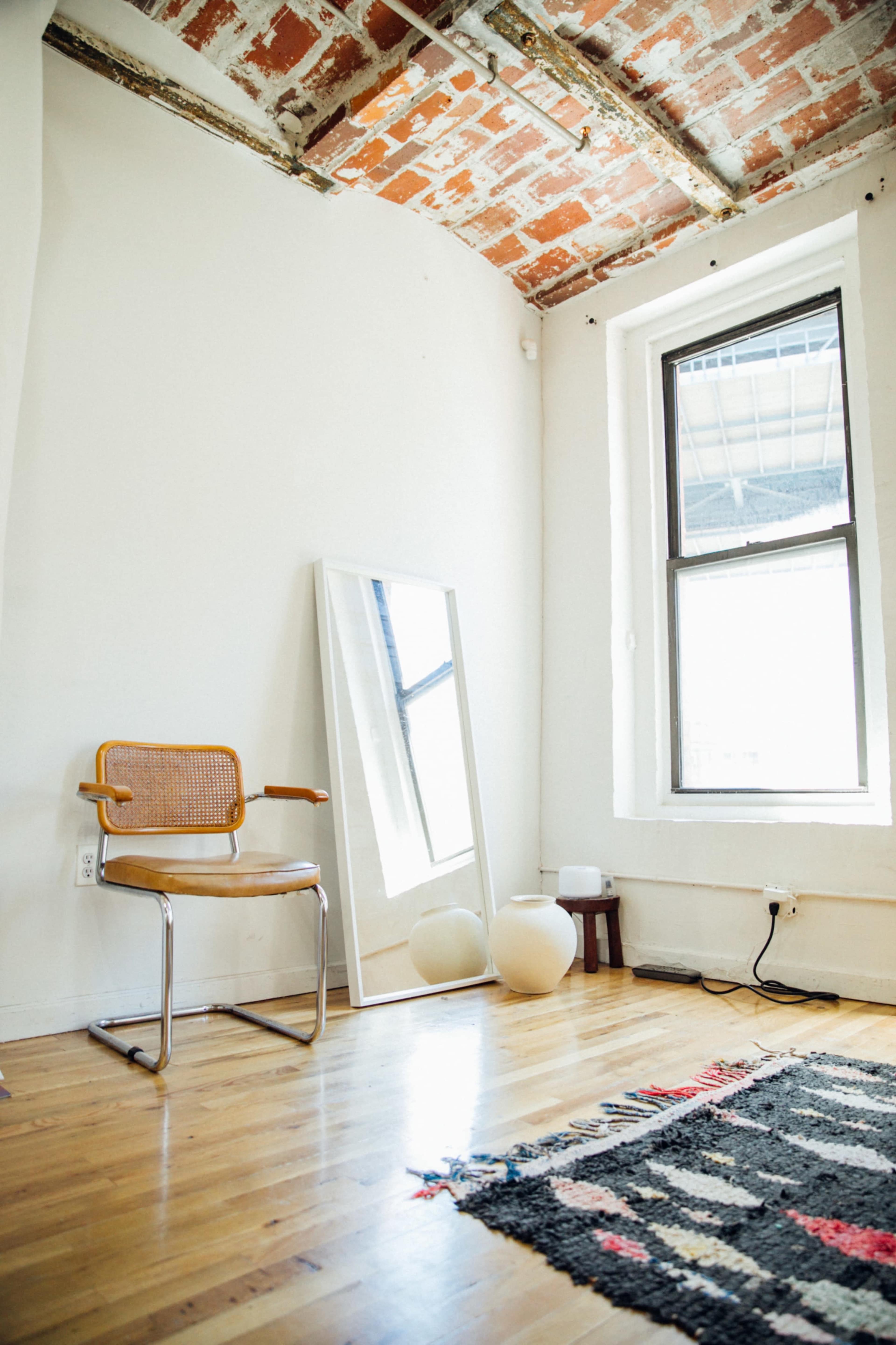 A minimalist room featuring a wooden floor, a chair, a large mirror, two round objects, and a window with exposed brick walls.