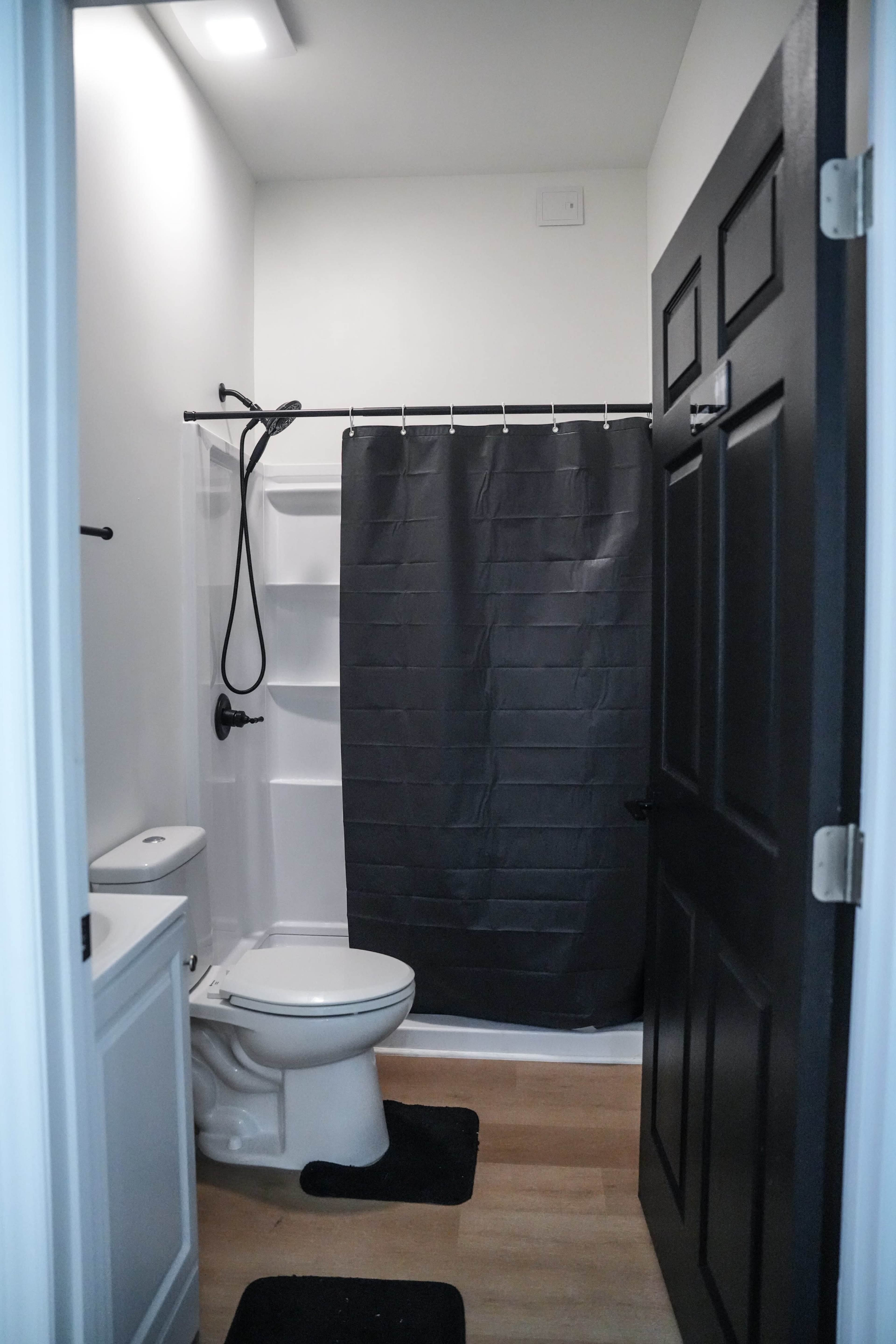 The image shows a small bathroom featuring a toilet, a shower with a dark curtain, and a door painted black.