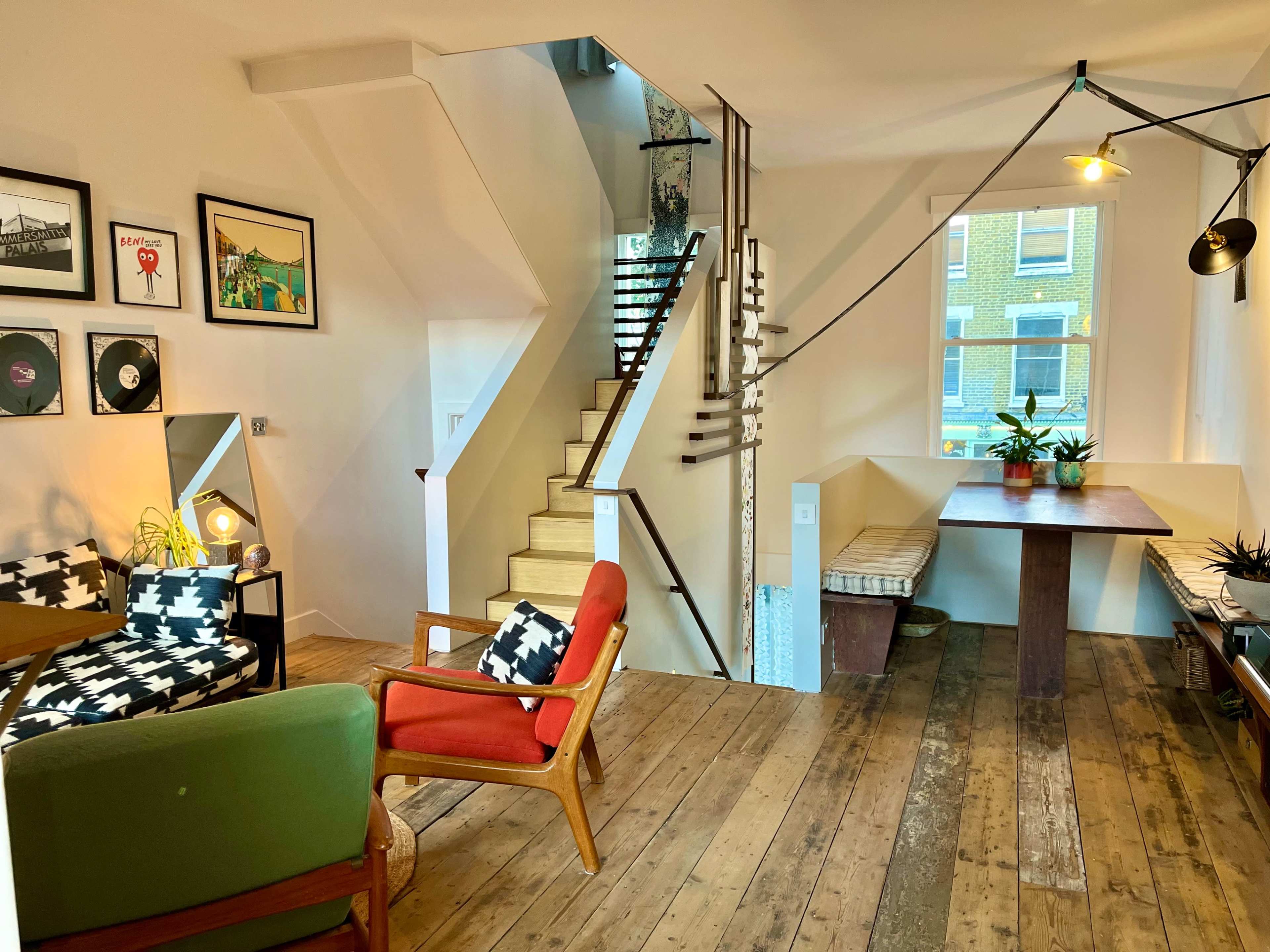 The image shows a modern interior space with wooden flooring, a staircase leading to an upper level, a dining table, and a seating area featuring patterned chairs and framed artwork.