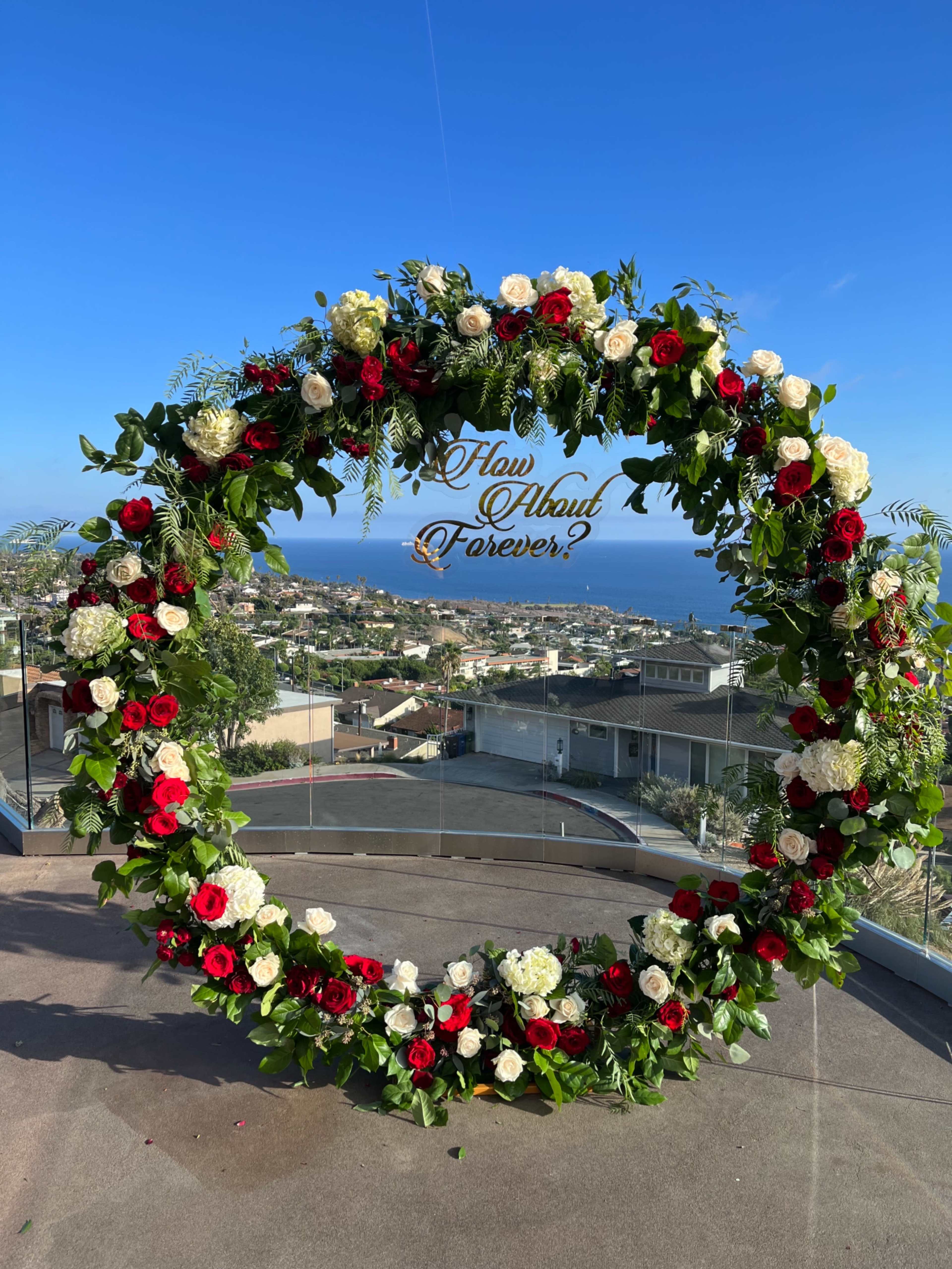 A large floral arch adorned with red and white roses is set against a backdrop of a coastal landscape.