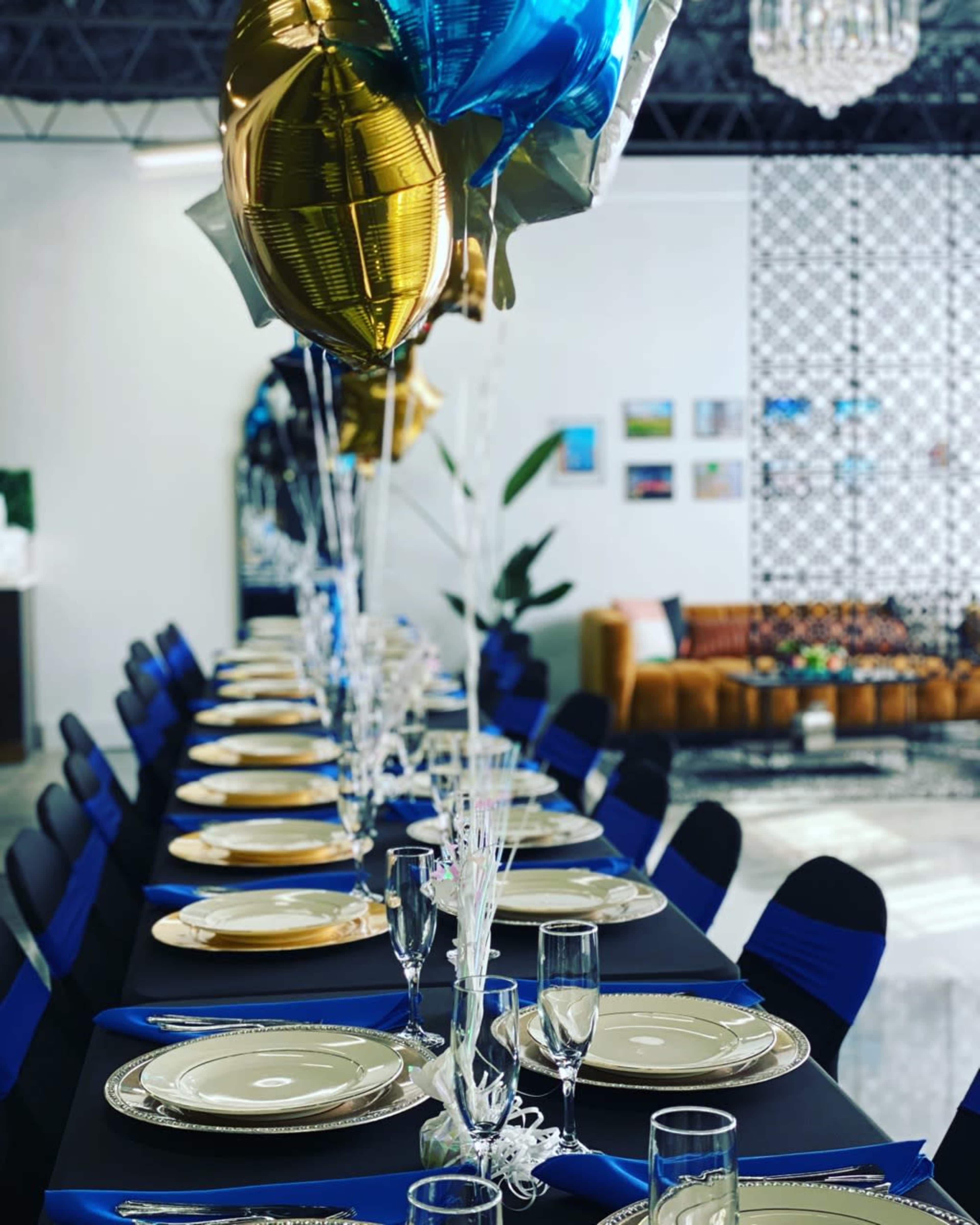 A long banquet table is set with plates and glassware, adorned with blue and gold balloons, against a backdrop of decorated walls and a couch.