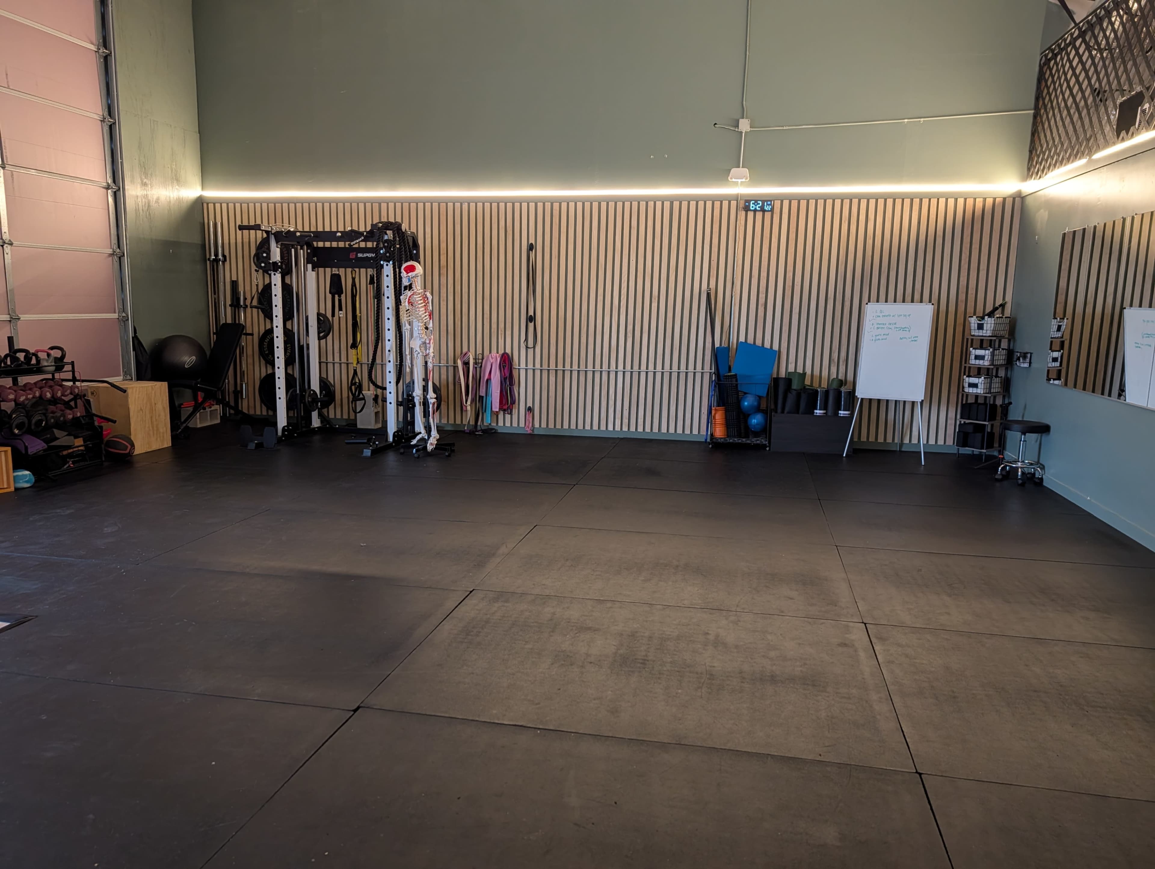 The image shows a spacious gym interior with exercise equipment, including a cable machine, free weights, and mats, against a wall with wooden paneling.