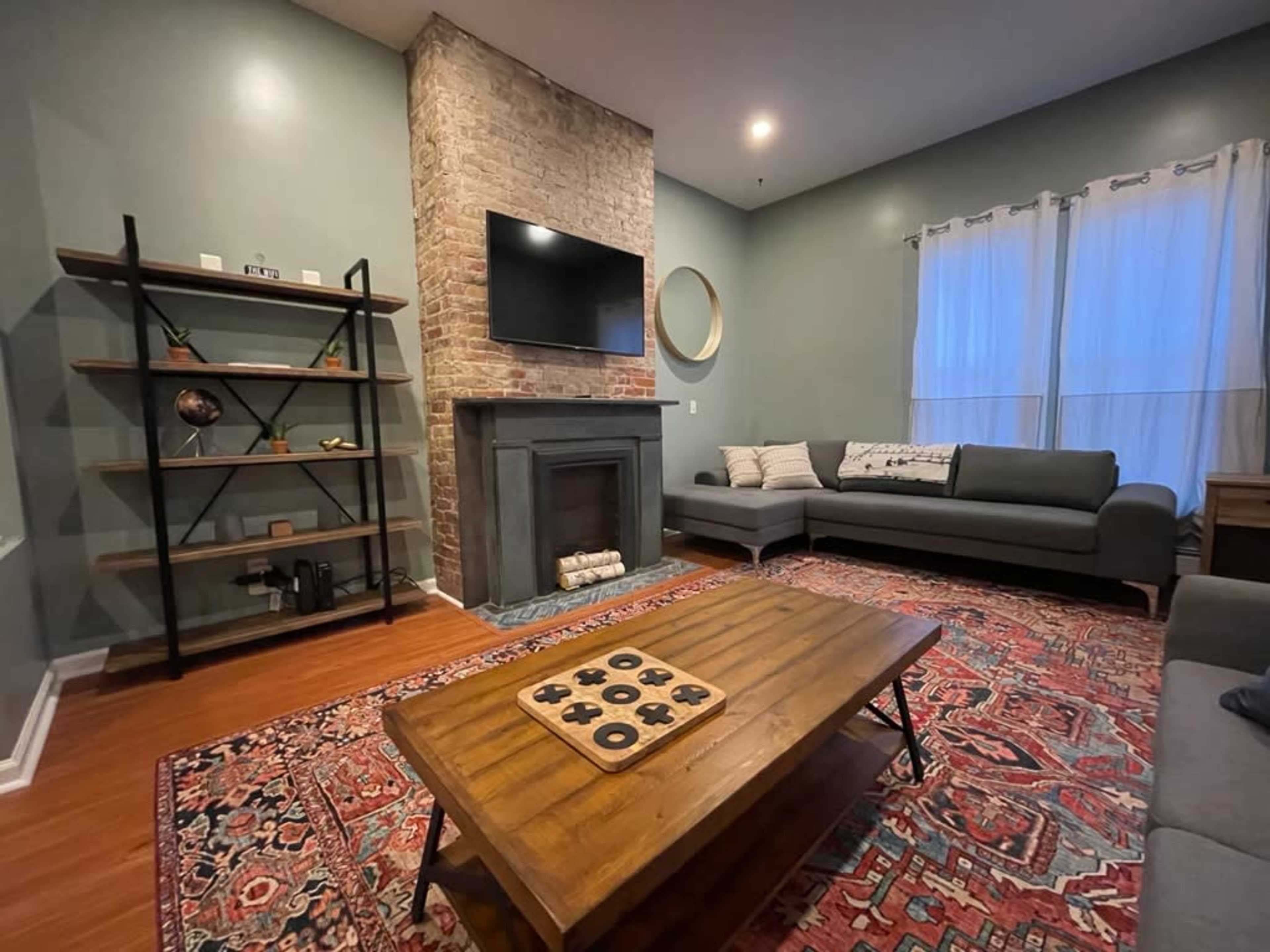 A cozy living room featuring a brick fireplace, a gray sectional sofa, a wooden coffee table with decorative items, and a large wall-mounted TV.