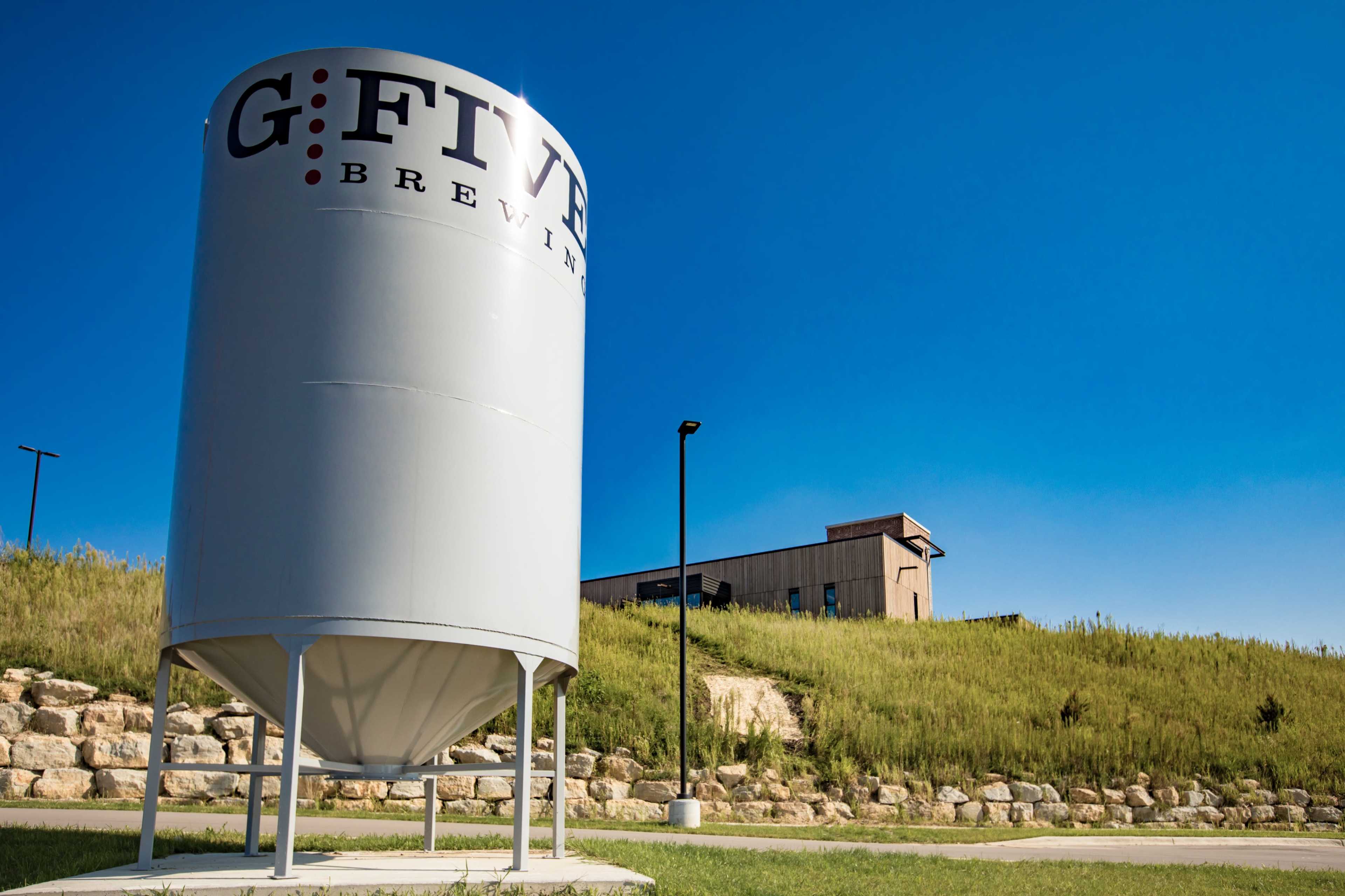 A large gray silo with the name "G.Five Brewing" stands in front of a green hillside and a building under a clear blue sky.
