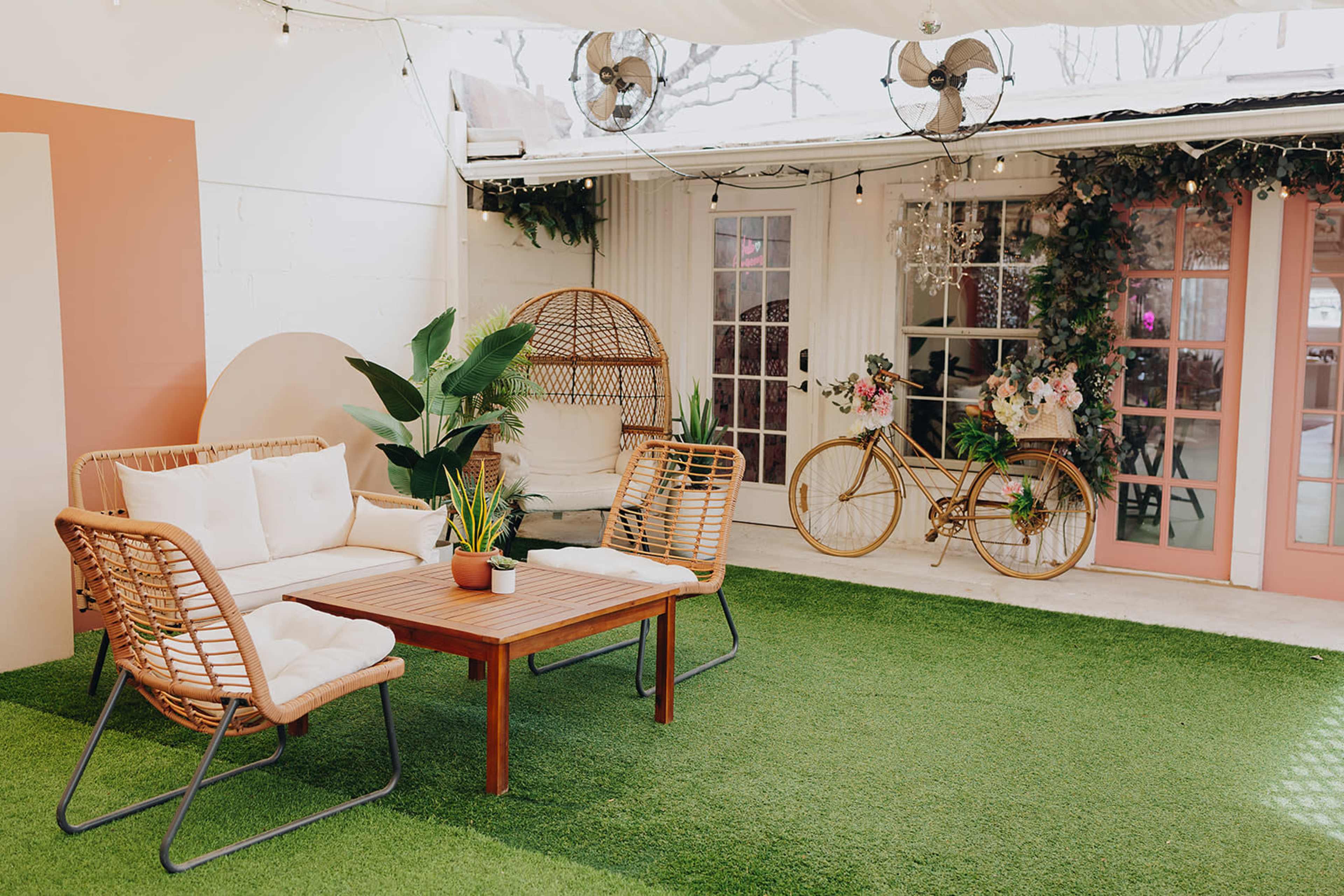 The image shows a cozy outdoor seating area with rattan furniture, a wooden coffee table, and decorative plants surrounded by white walls and doors with pink accents.