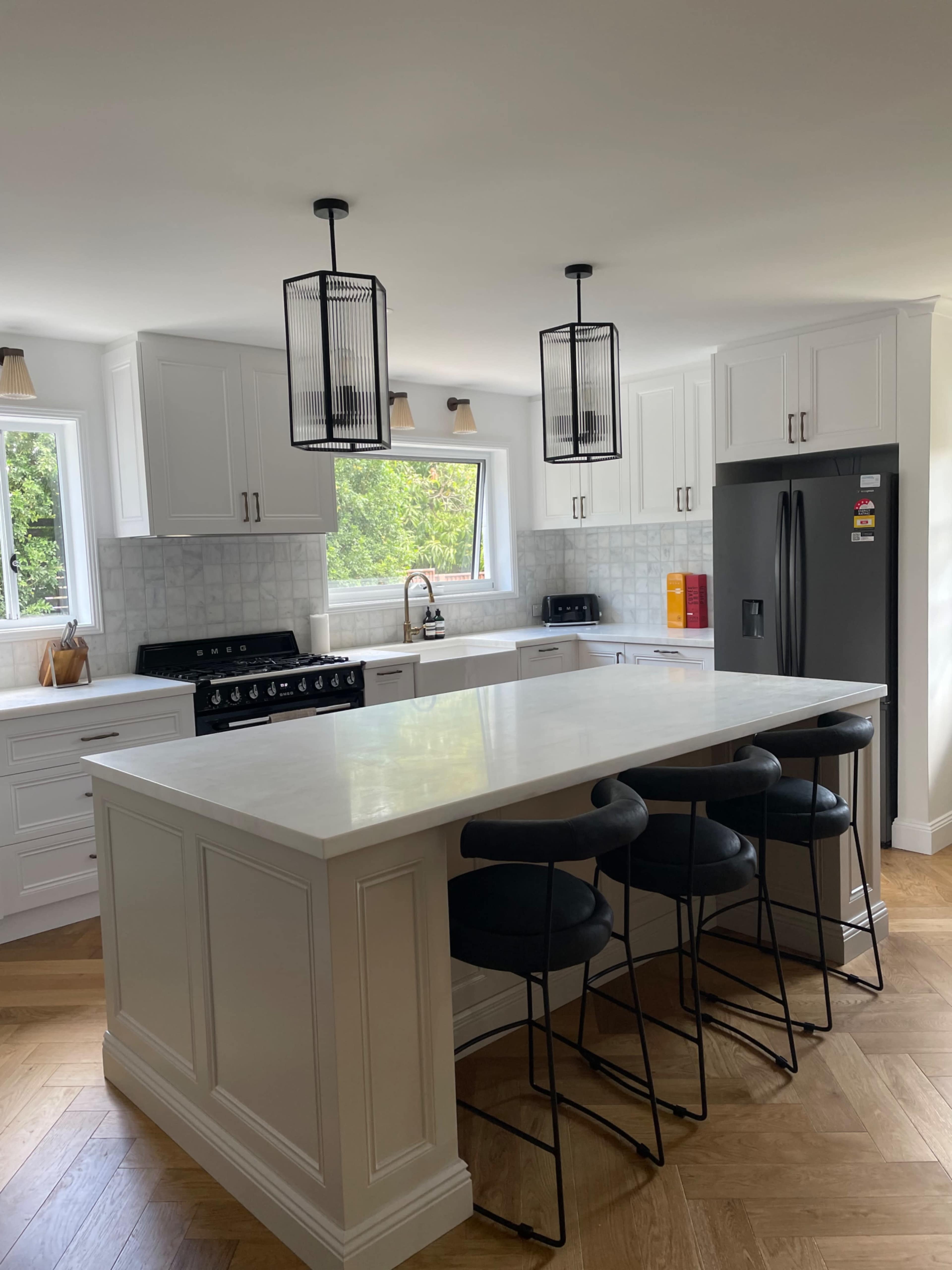 A modern kitchen features a large island with four black stools, white cabinetry, a stainless steel refrigerator, and pendant lighting.