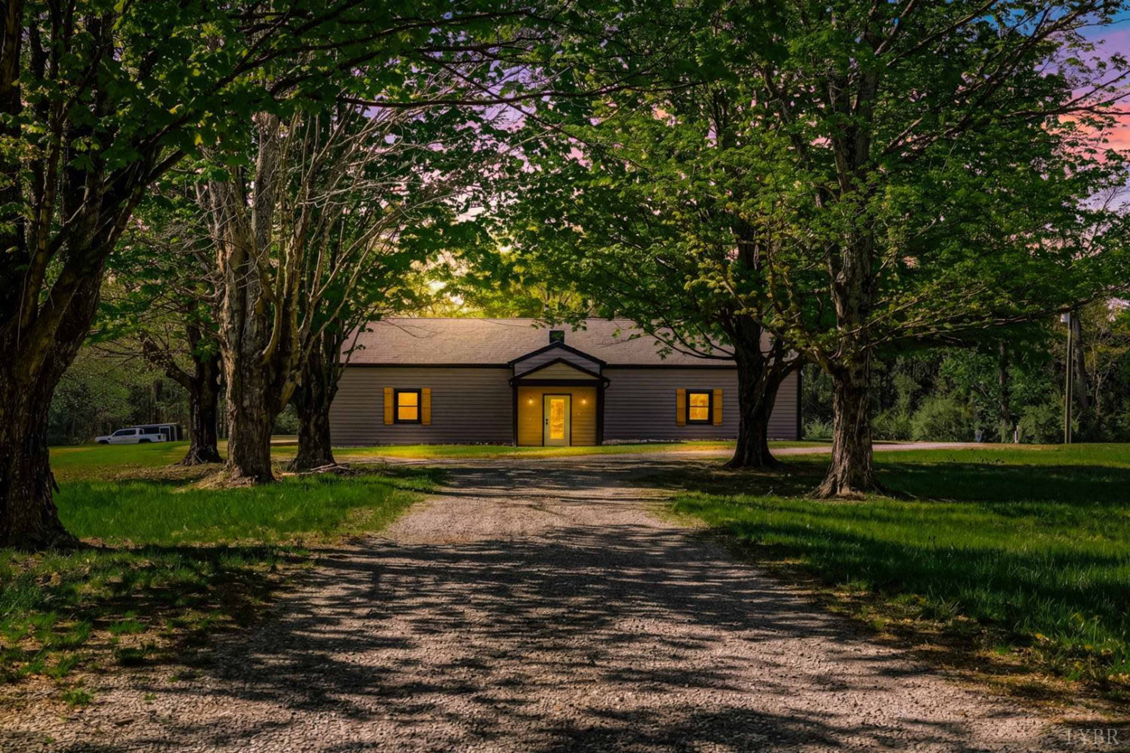 A gravel driveway leads to a house surrounded by trees, with lit windows and a sunset in the background.