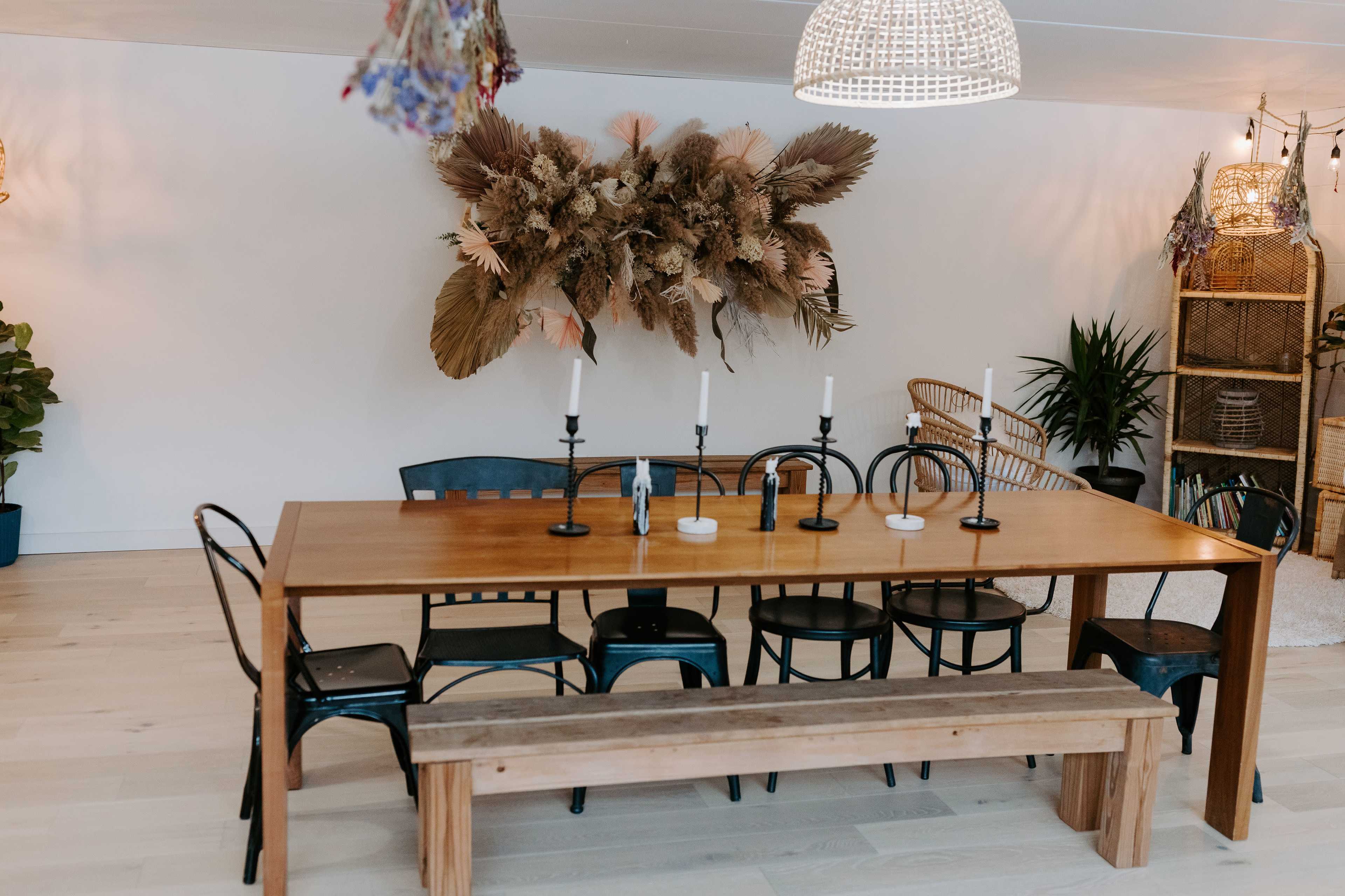 A wooden dining table with black chairs and a bench is set in a well-lit room featuring a decorative wall arrangement of dried plants.