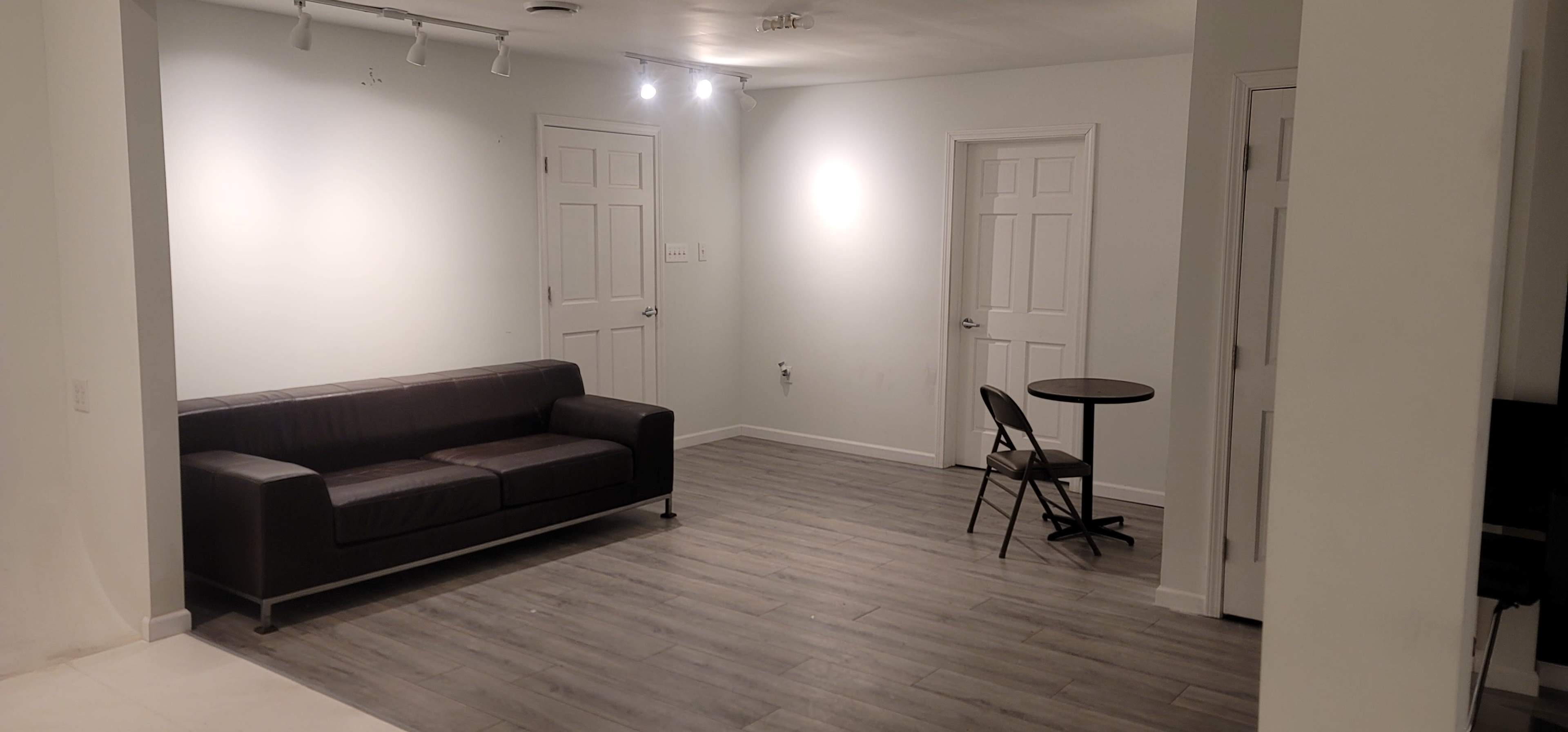 The room features a dark couch, a small table with a chair, and two white doors on opposite walls.