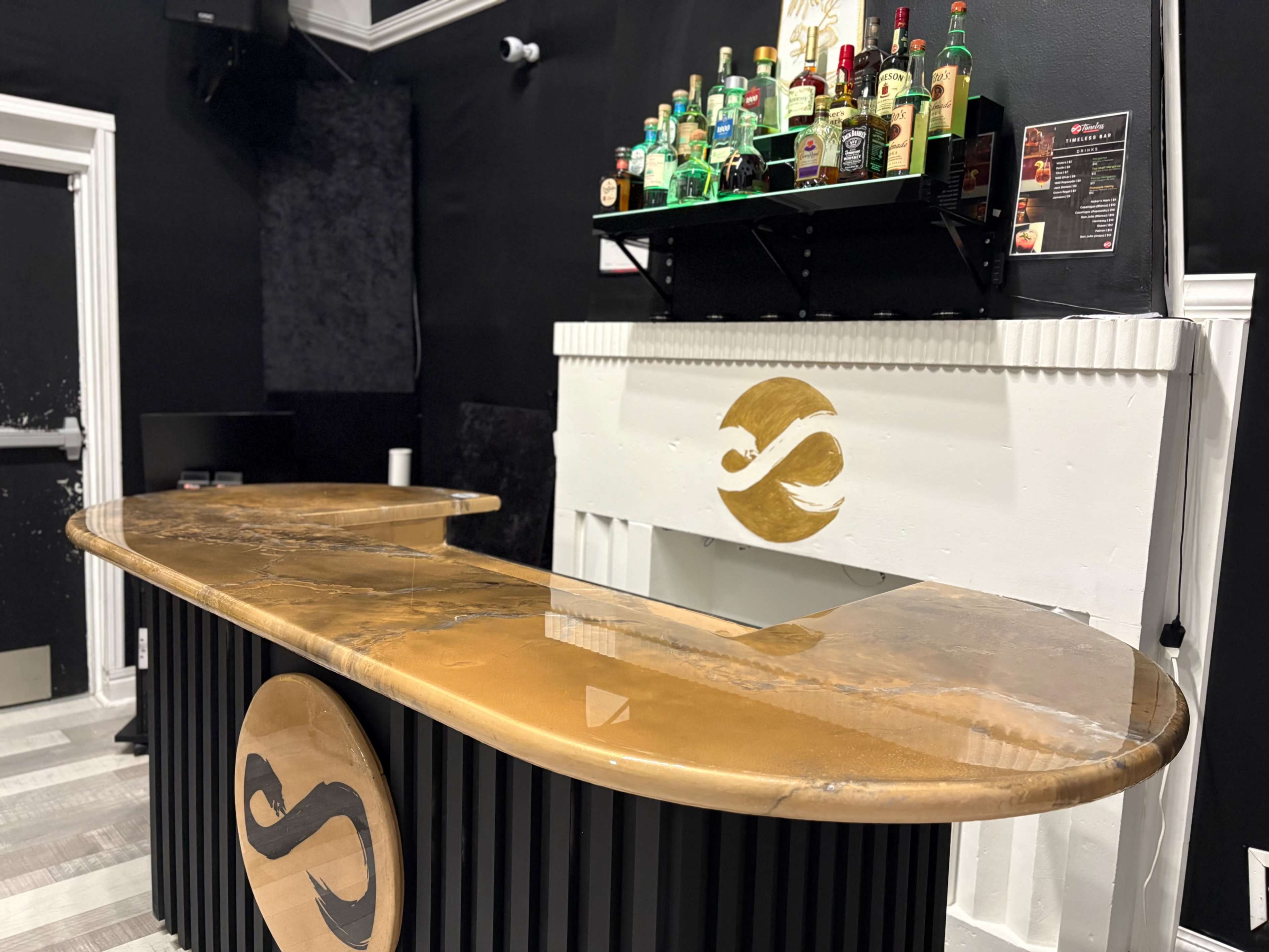 The image shows a modern bar with a glossy, golden countertop, a black backdrop, and shelves stocked with various bottles of liquor.