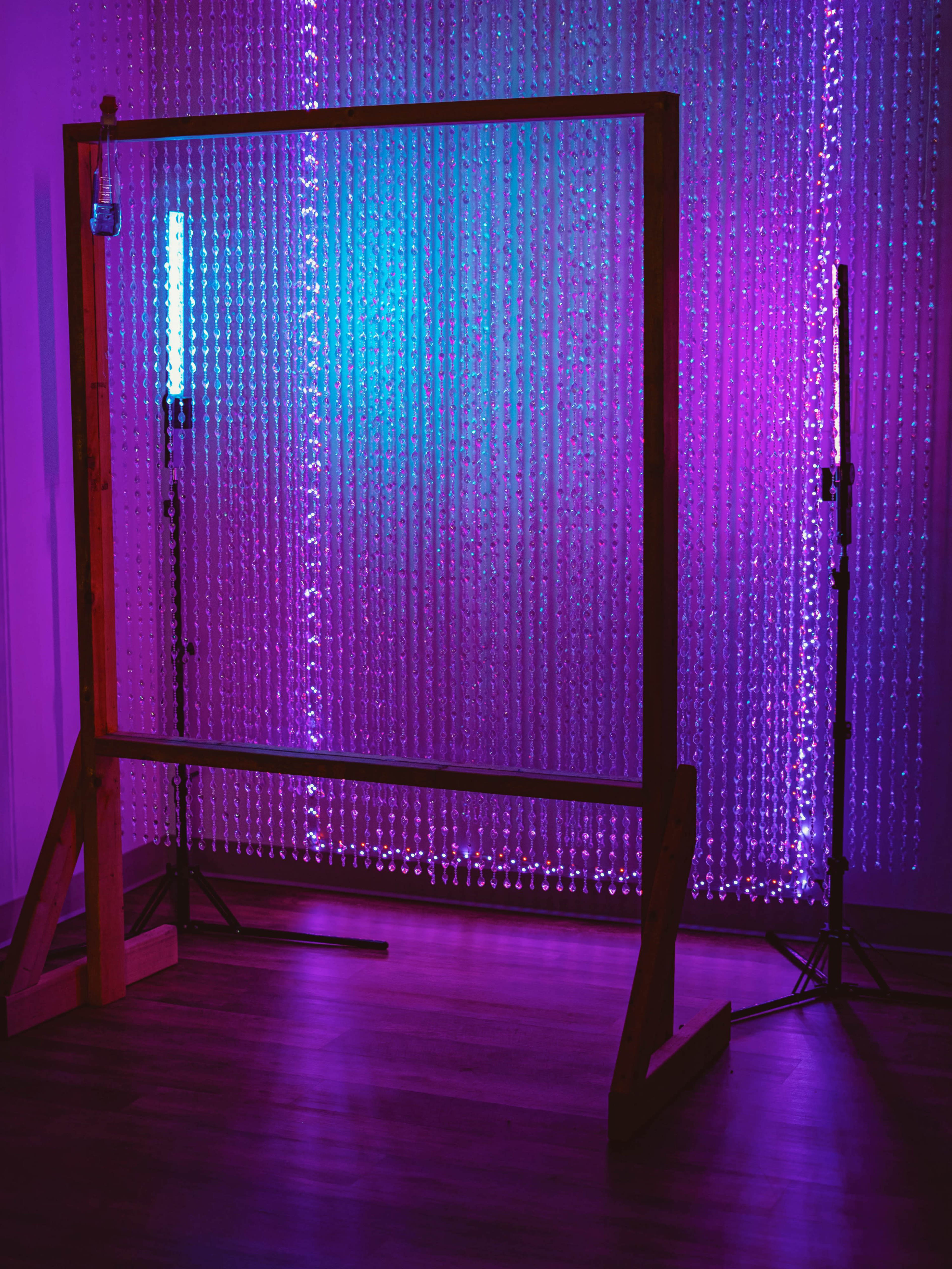 The image features a wooden frame positioned against a backdrop of hanging beads illuminated by pink and blue lights.