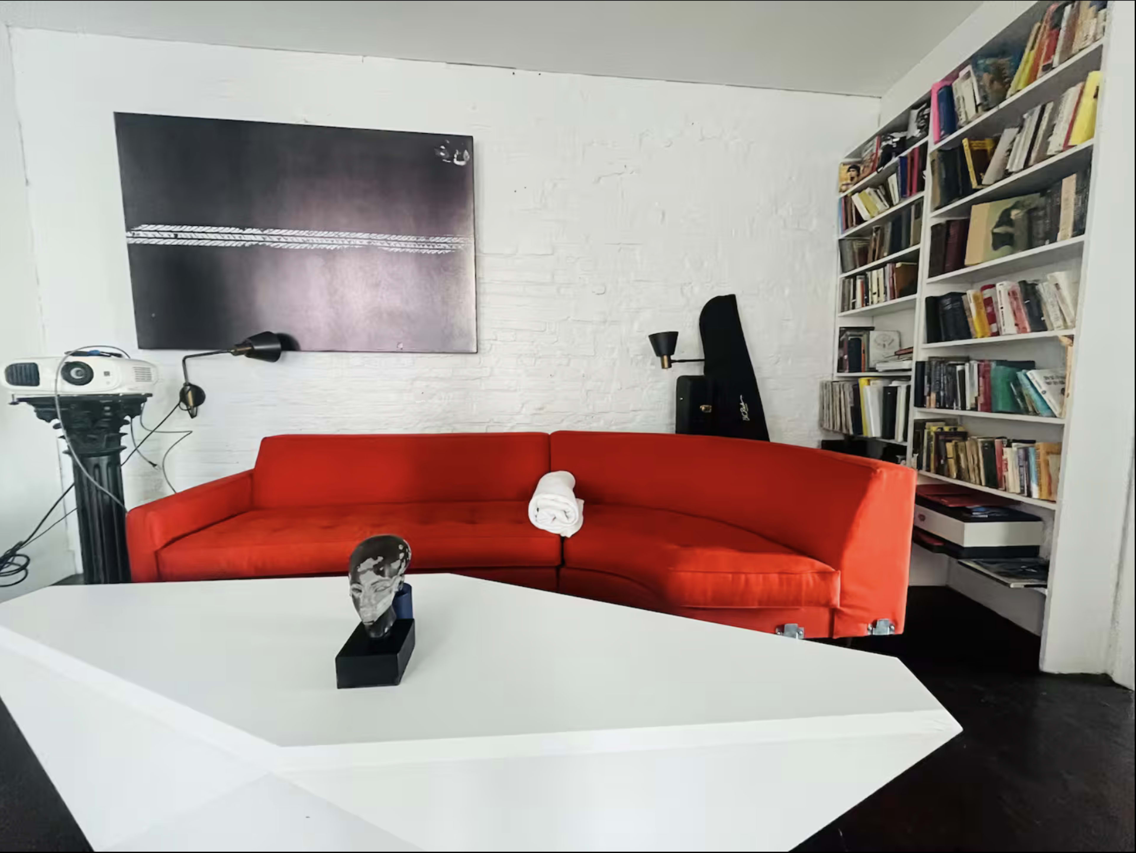 The image shows a cozy living space featuring a red curved sofa, a white coffee table, a projector, and a bookshelf filled with books against a white brick wall.