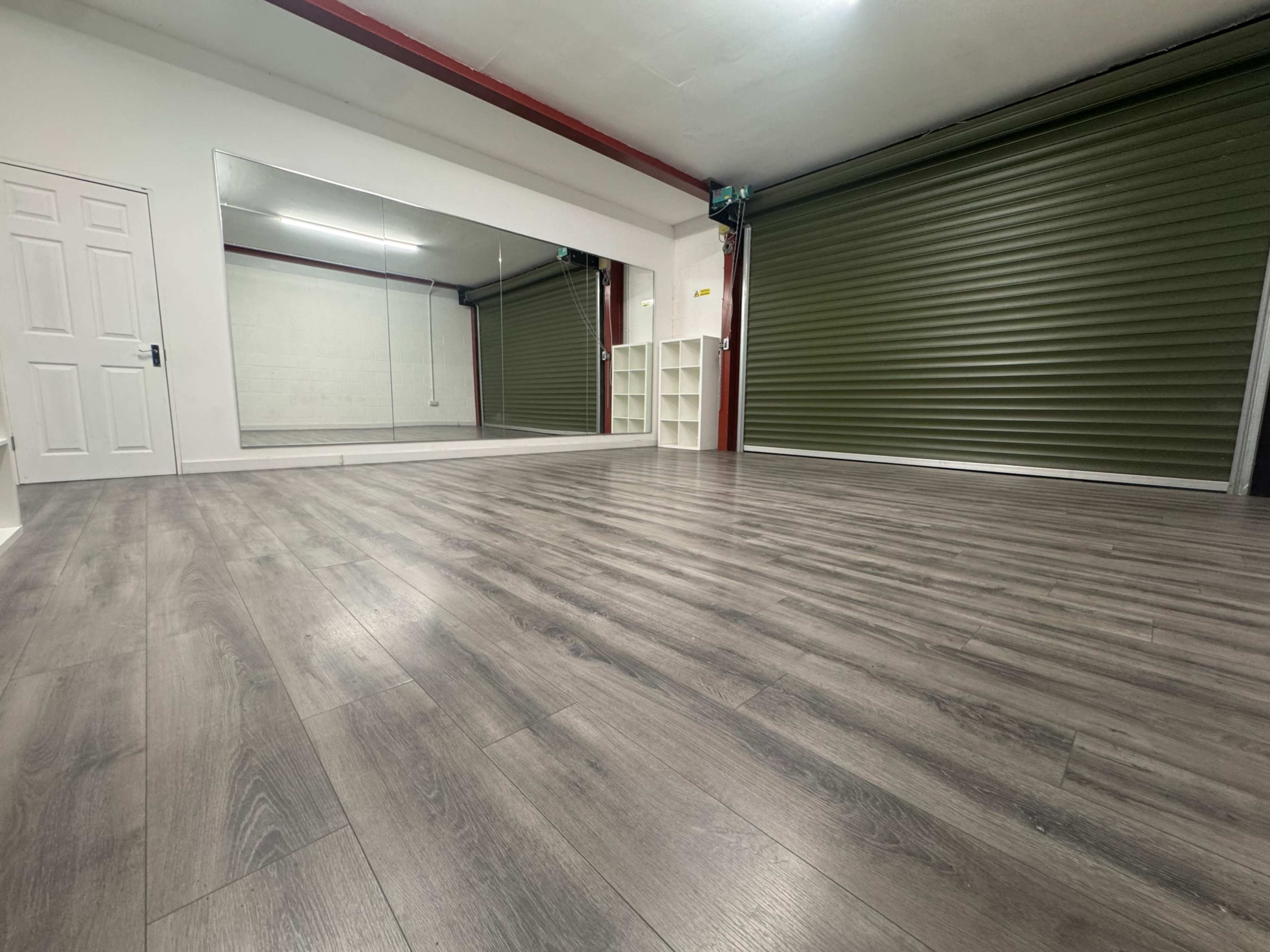 The image shows a spacious indoor room with wooden flooring, a large mirror on one wall, and green rolling shutters closed on another side.