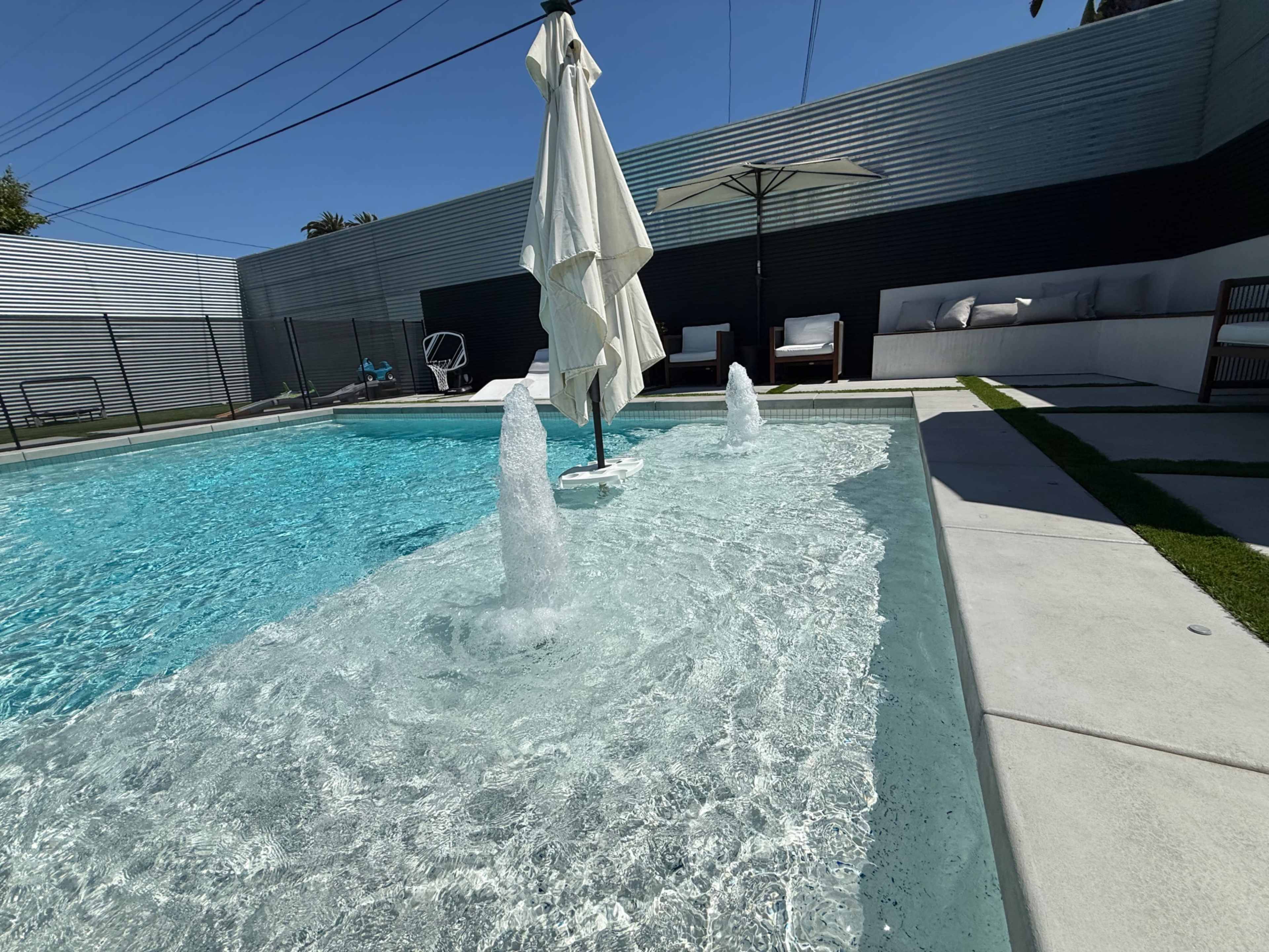 The image shows a modern swimming pool with water fountains, surrounded by lounge chairs and an umbrella, set against a contemporary backdrop.