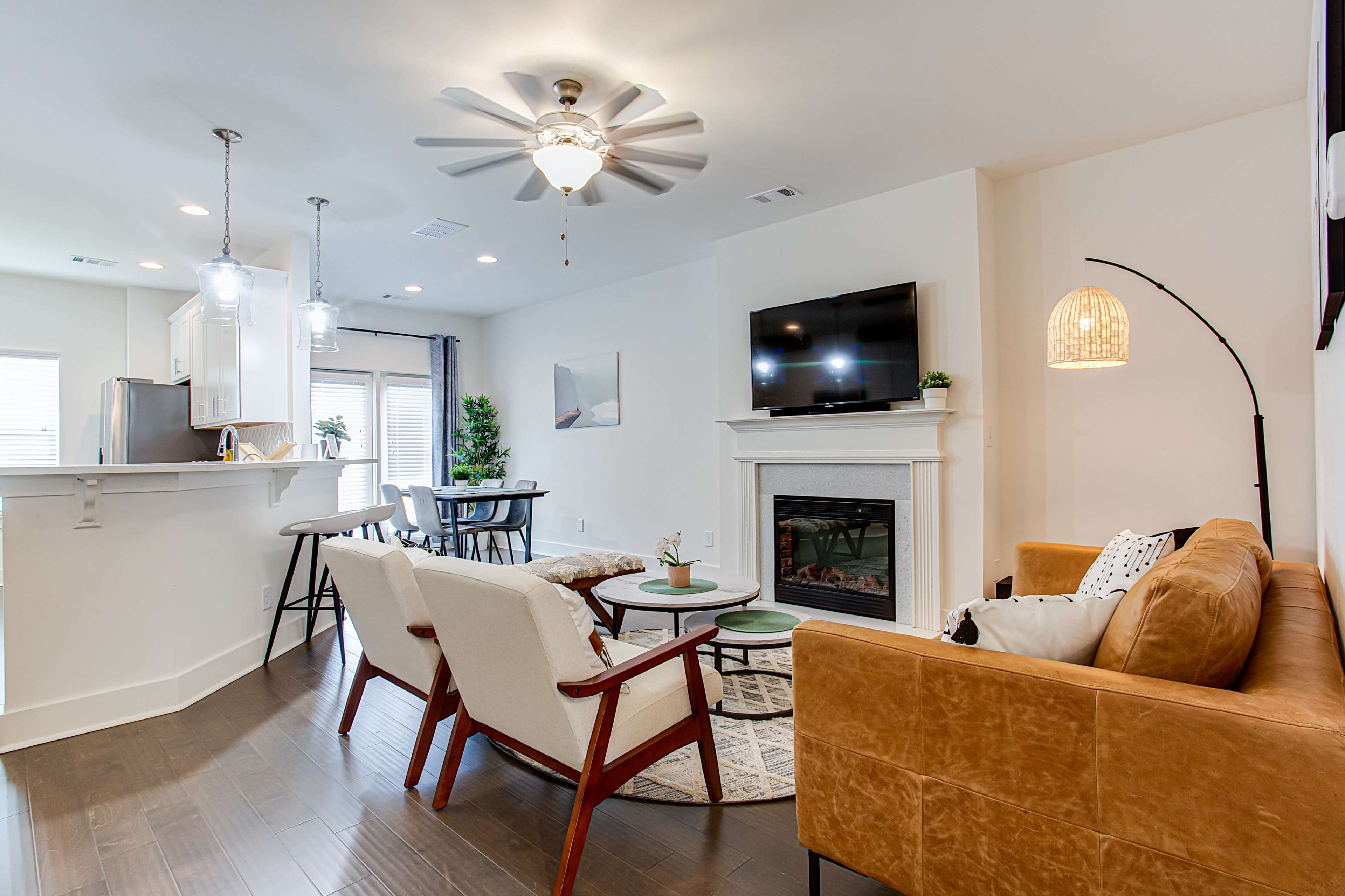 A modern living room with a couch, chairs, a fireplace, and a television, accented by pendant lighting and a dining area in the background.