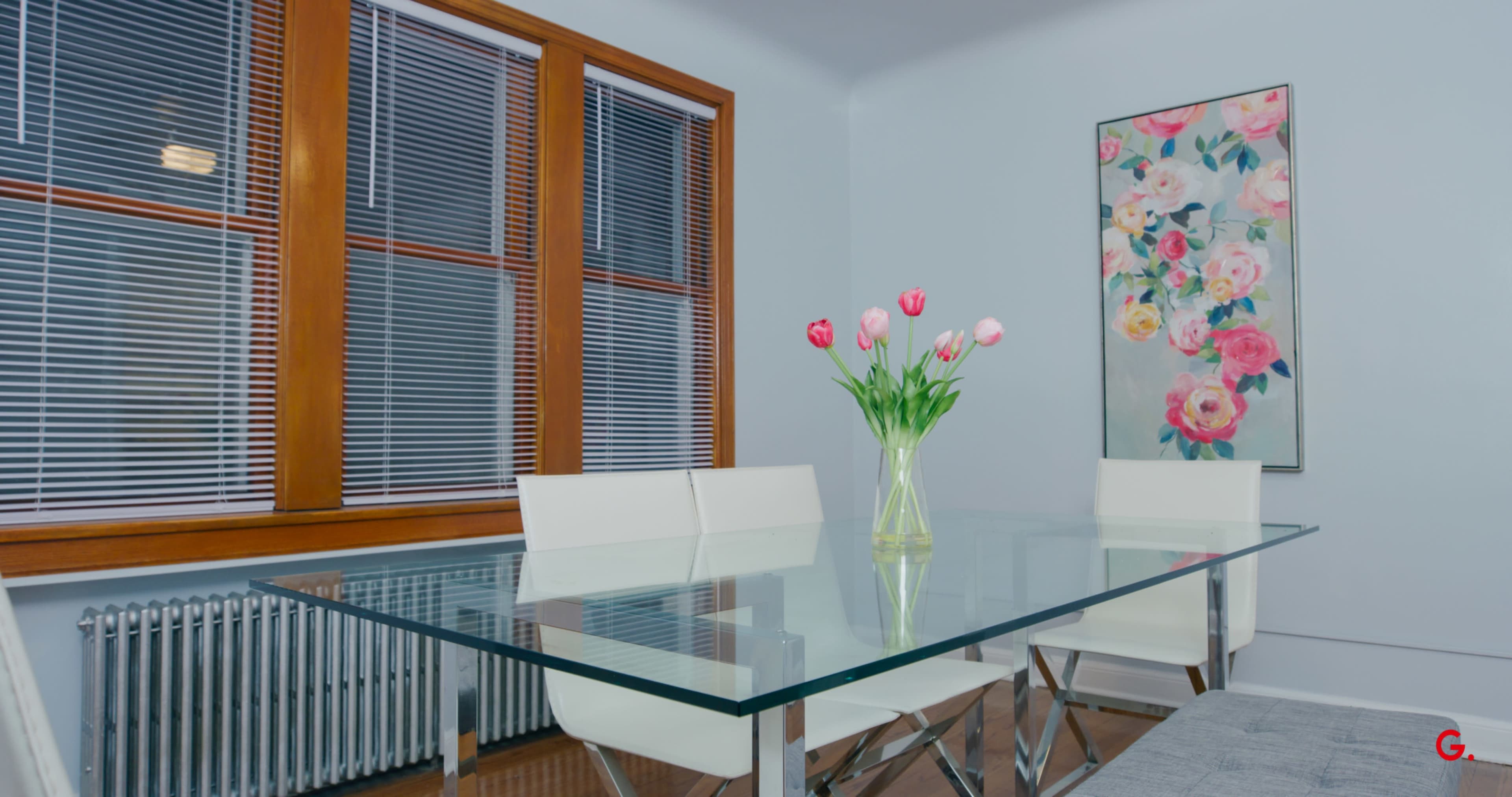 A glass dining table with white chairs is set near a window, while a vase of tulips sits in the center alongside a floral artwork on the wall.