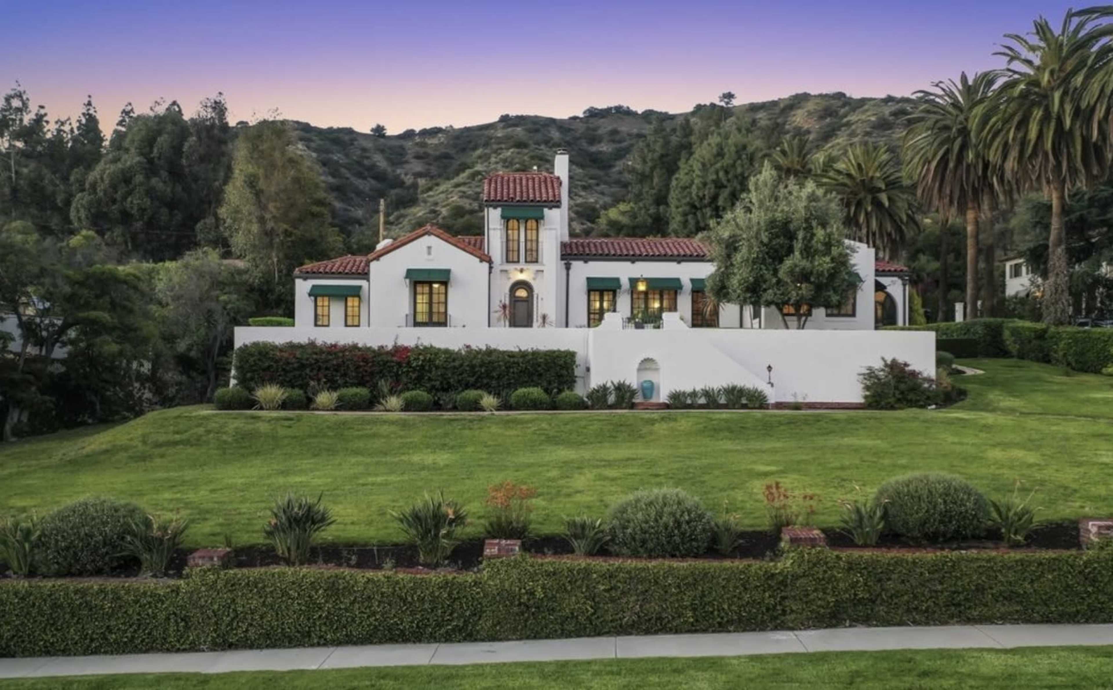 A white stucco house with a tile roof and green shutters is set on a manicured lawn surrounded by palm trees and shrubs in a hillside area.
