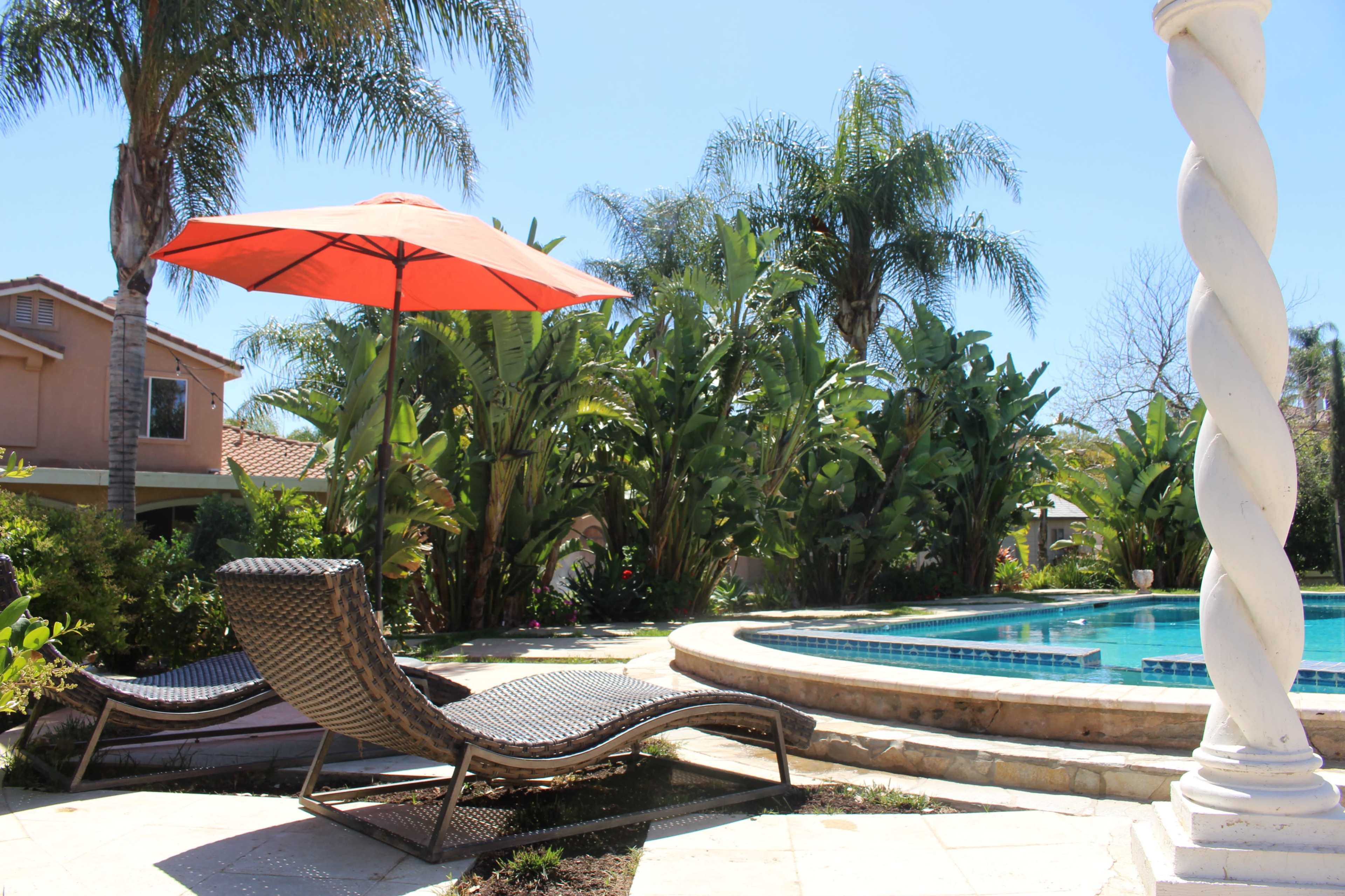 A lounge chair with an orange umbrella is positioned beside a swimming pool surrounded by tropical plants.
