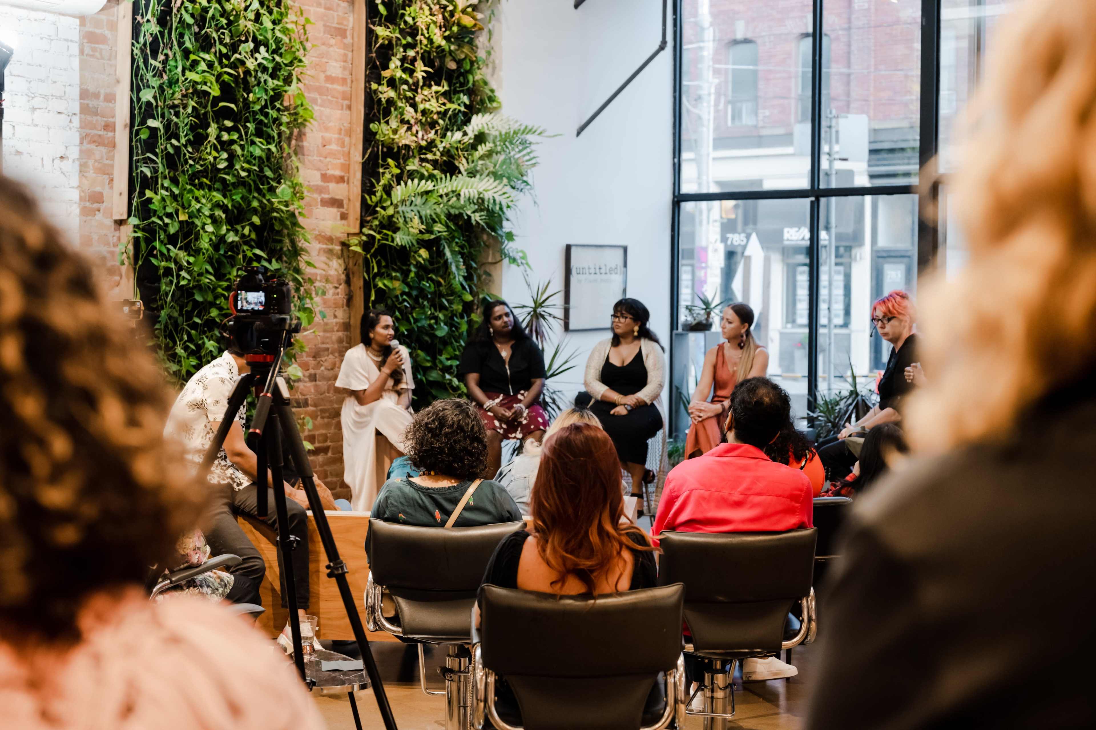 A group of six women participates in a panel discussion in a spacious room with large windows and greenery on the walls, while an audience listens in chairs.