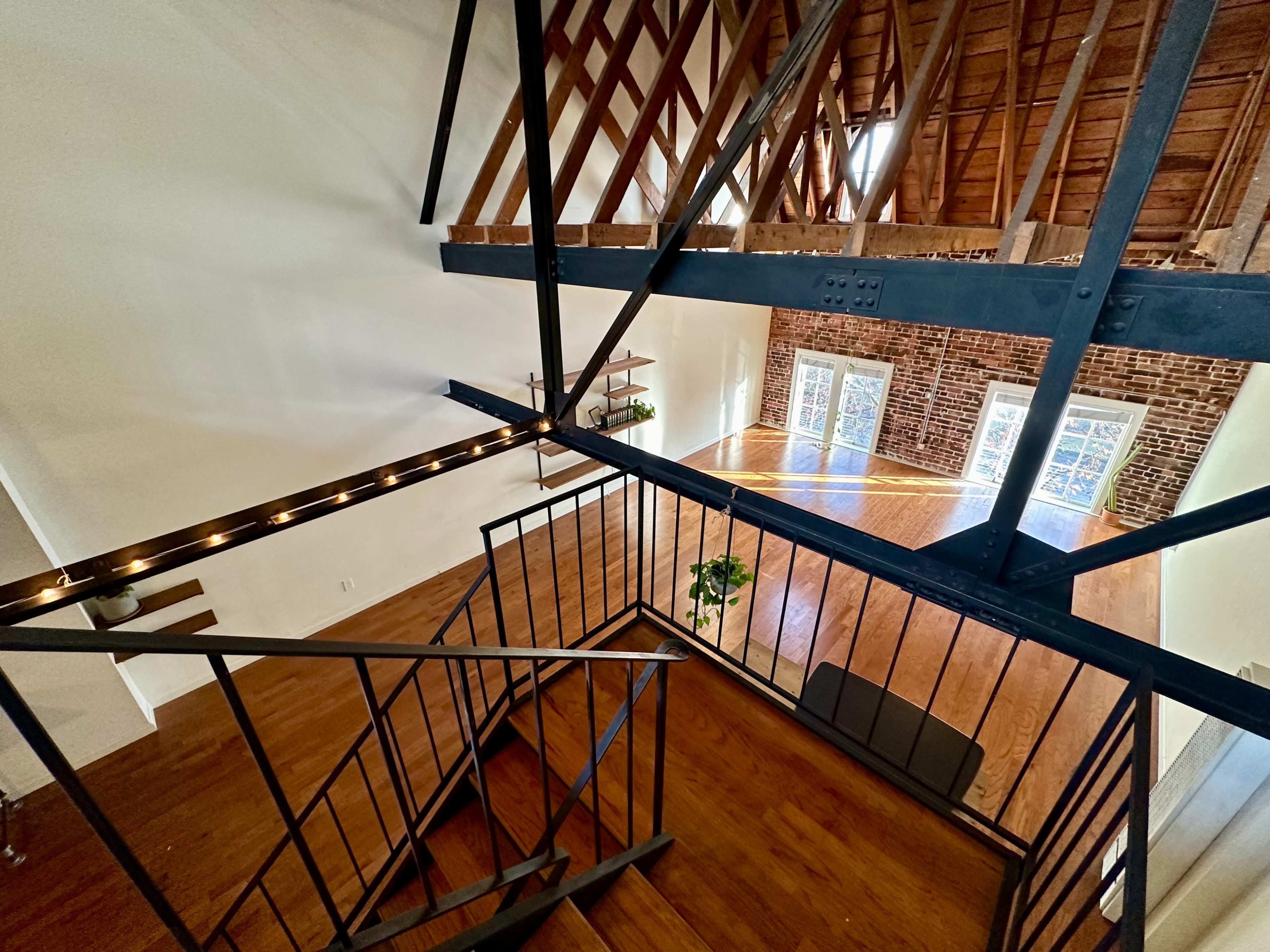 A staircase leads downward into an open space with exposed brick walls and wooden beams, featuring large windows that allow natural light to fill the area.