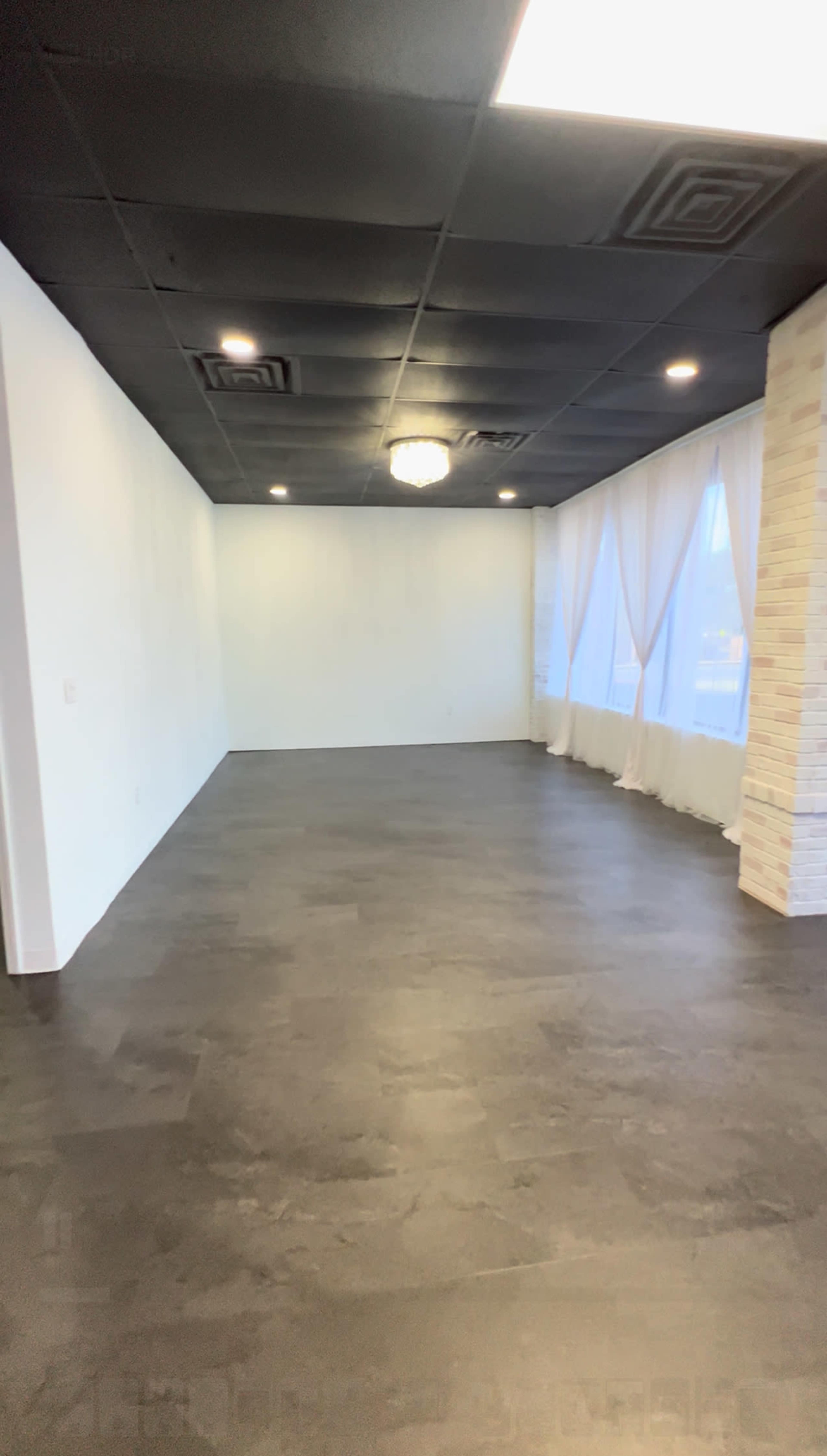 The image shows an empty room with dark flooring, white walls, and long sheer curtains allowing light to enter from large windows.