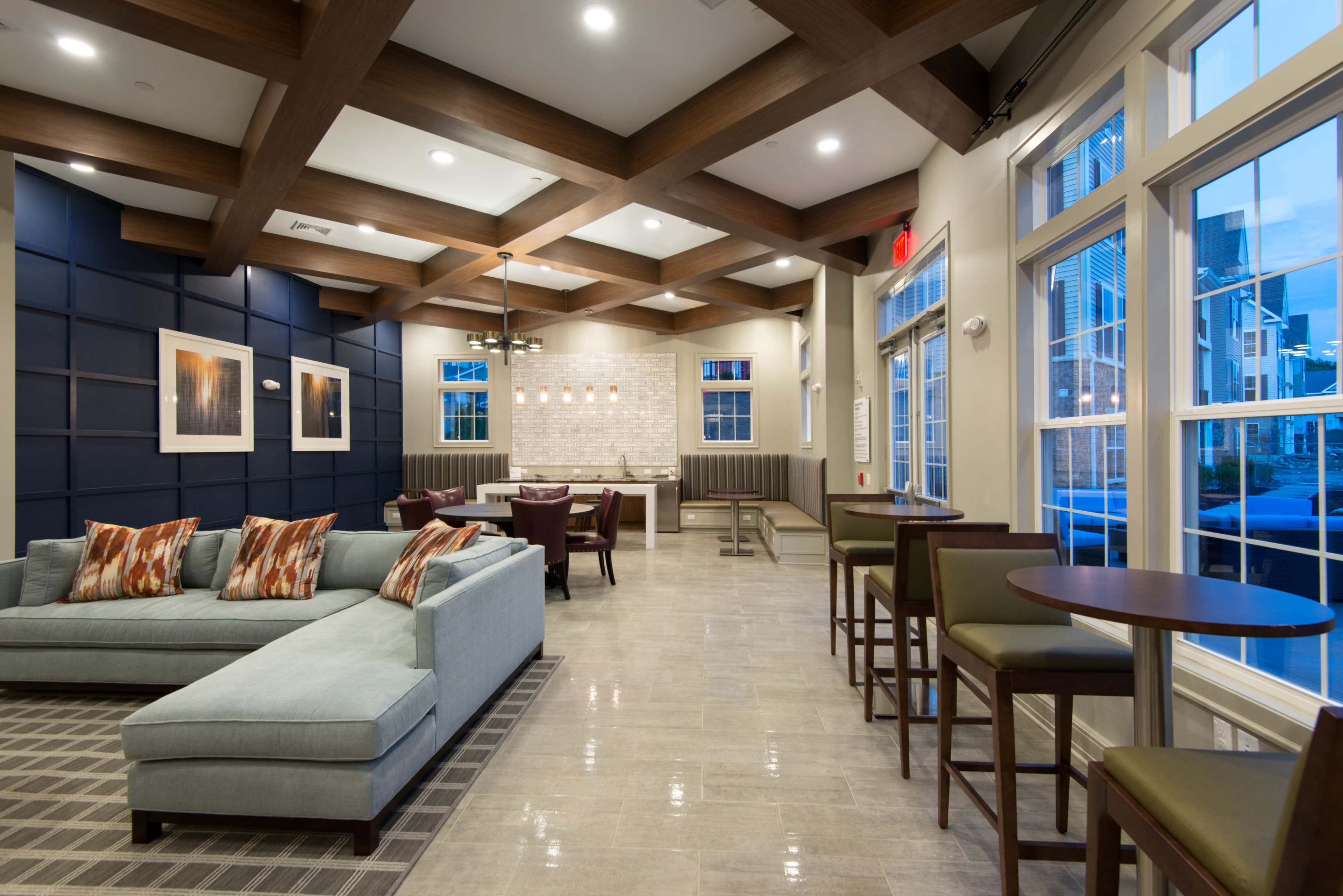 A modern common area with a blue sectional sofa, wooden beams on the ceiling, and a small dining space with high chairs and tables.