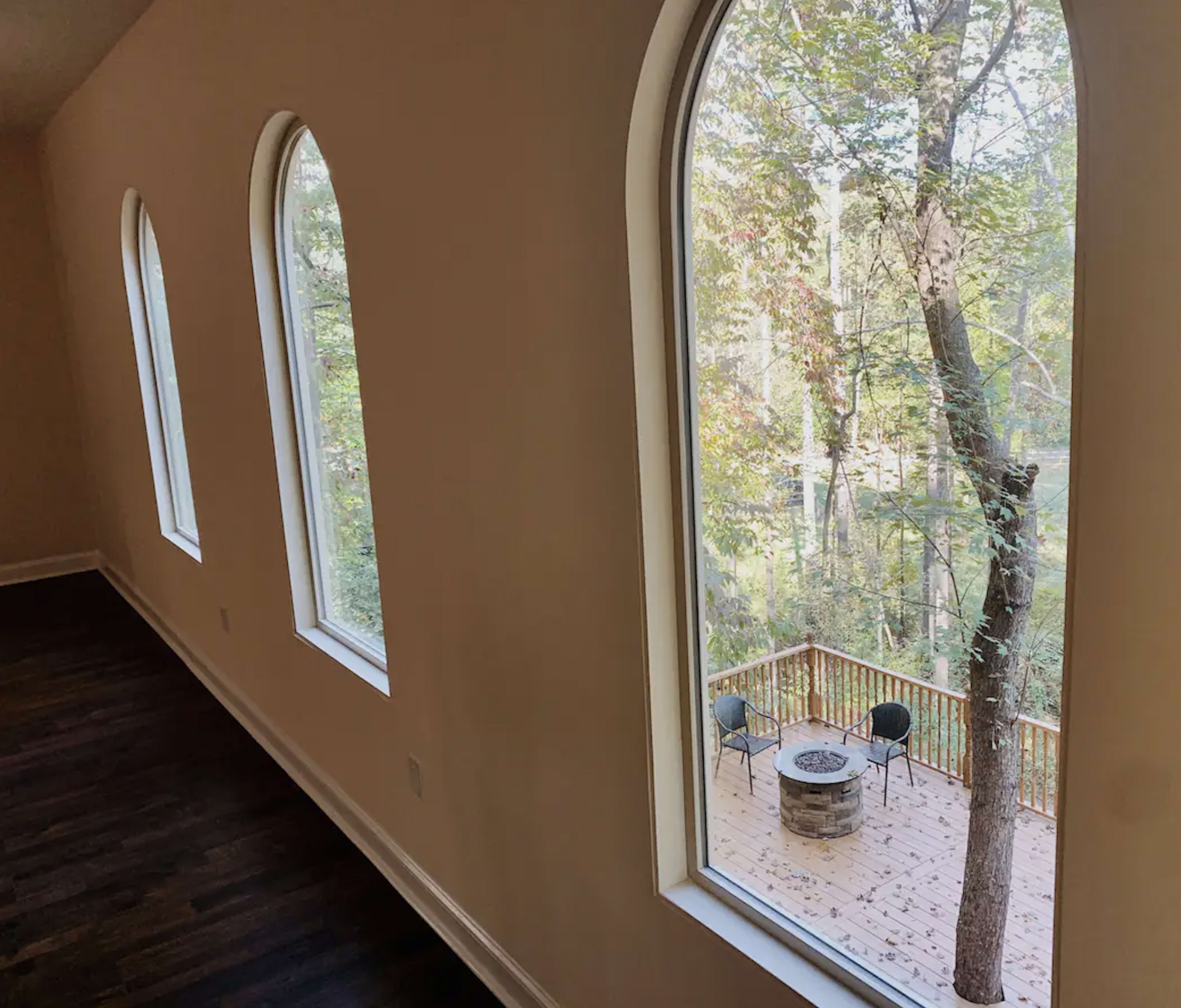 A room features large arched windows that overlook a wooden deck surrounded by trees.