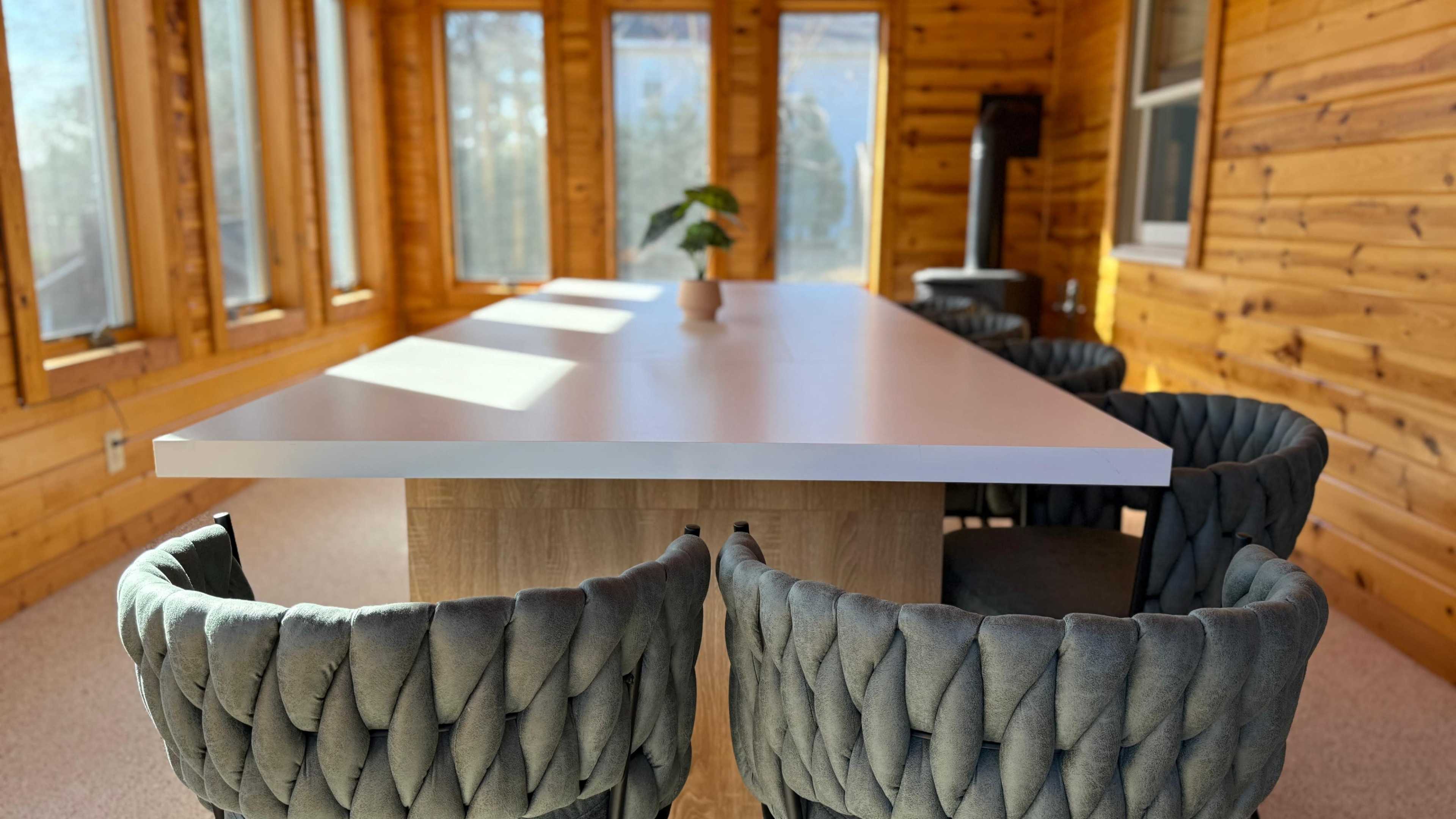 A modern table with a smooth white top is surrounded by textured gray chairs in a sunlit wood-paneled room with large windows.