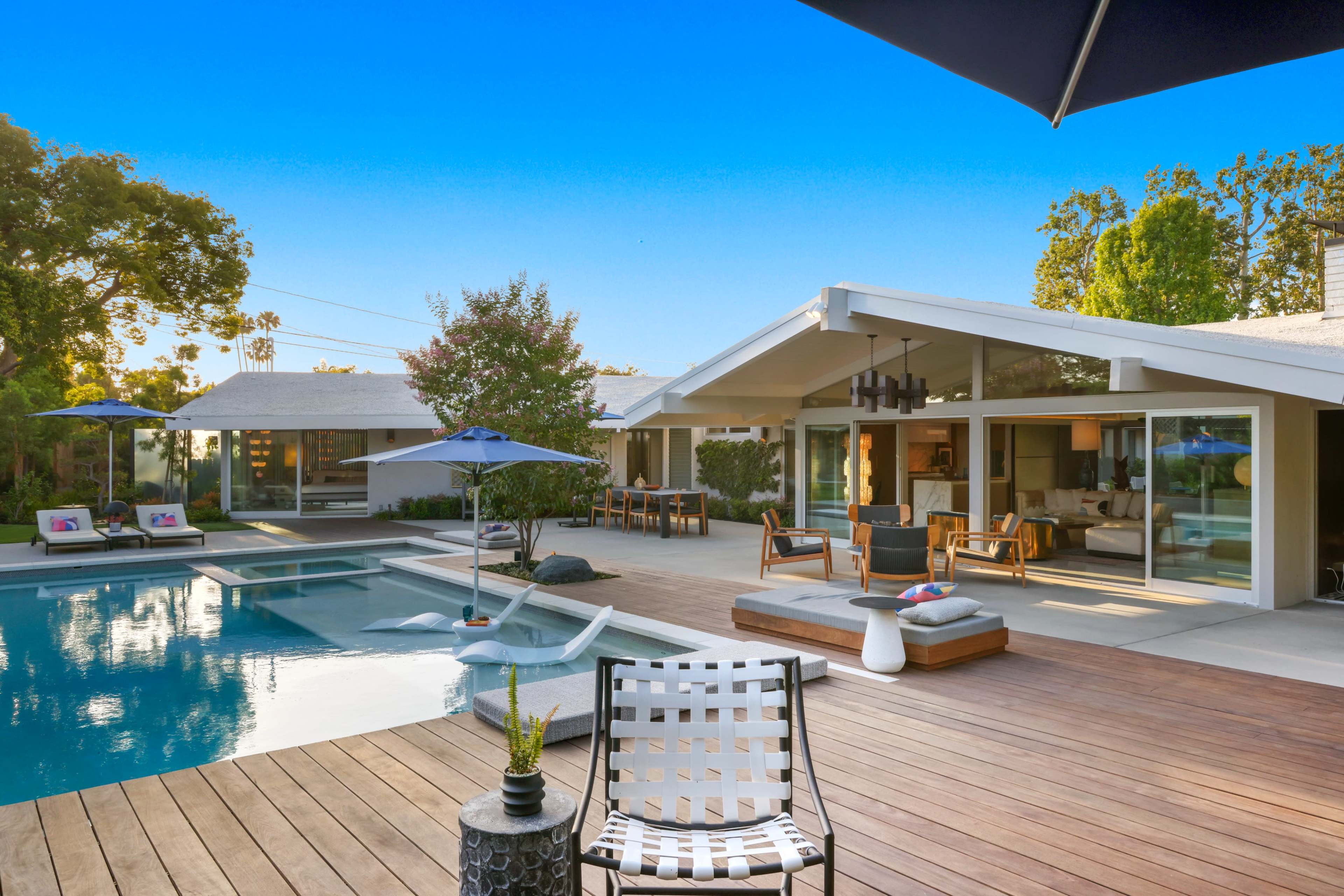 A modern backyard with a swimming pool, wooden decking, lounge chairs, and a house featuring large glass doors and an outdoor seating area.