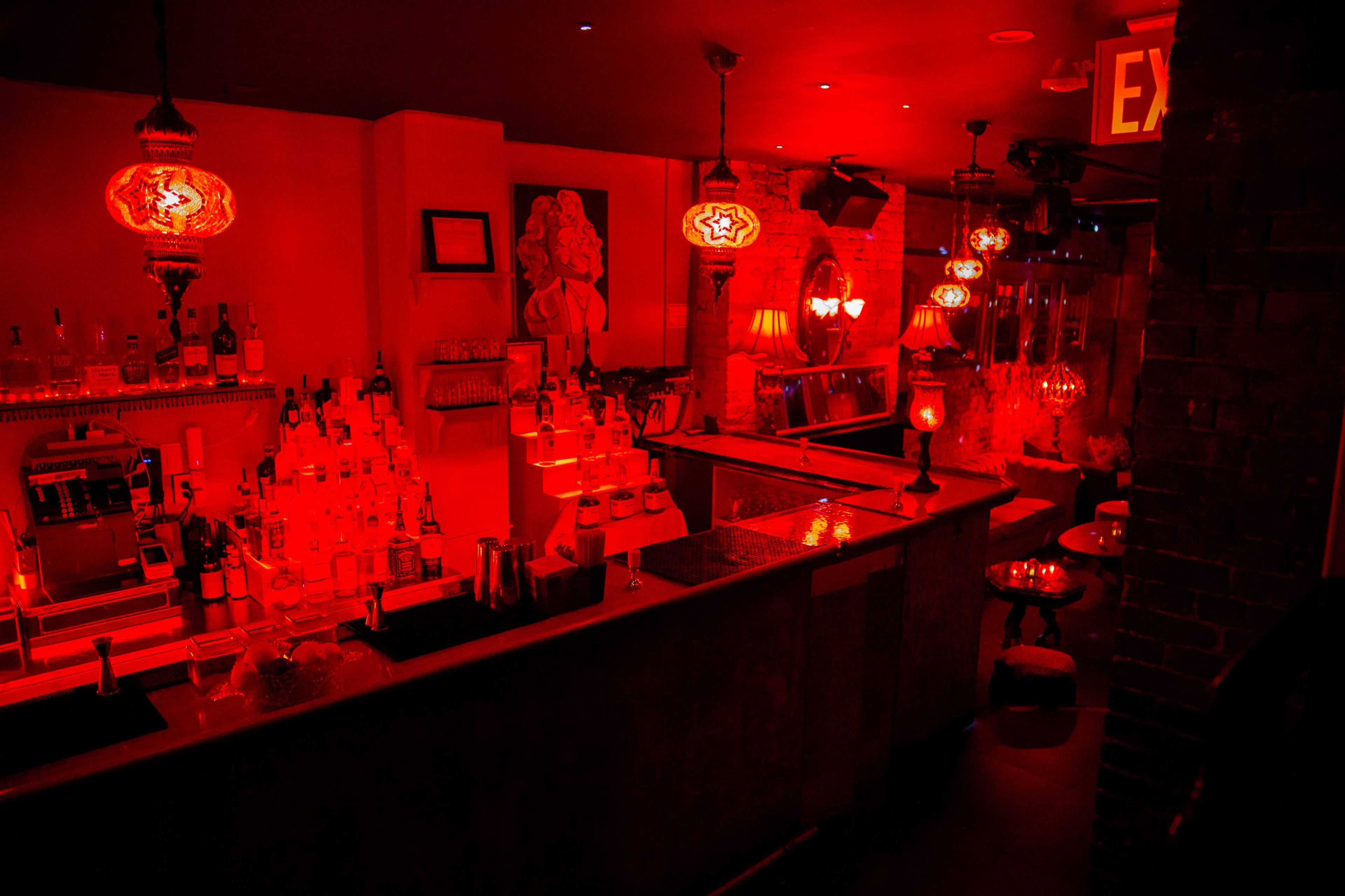 The image shows a dimly lit bar with red lighting, featuring a long counter stocked with various bottles and decorative pendant lamps hanging from the ceiling.