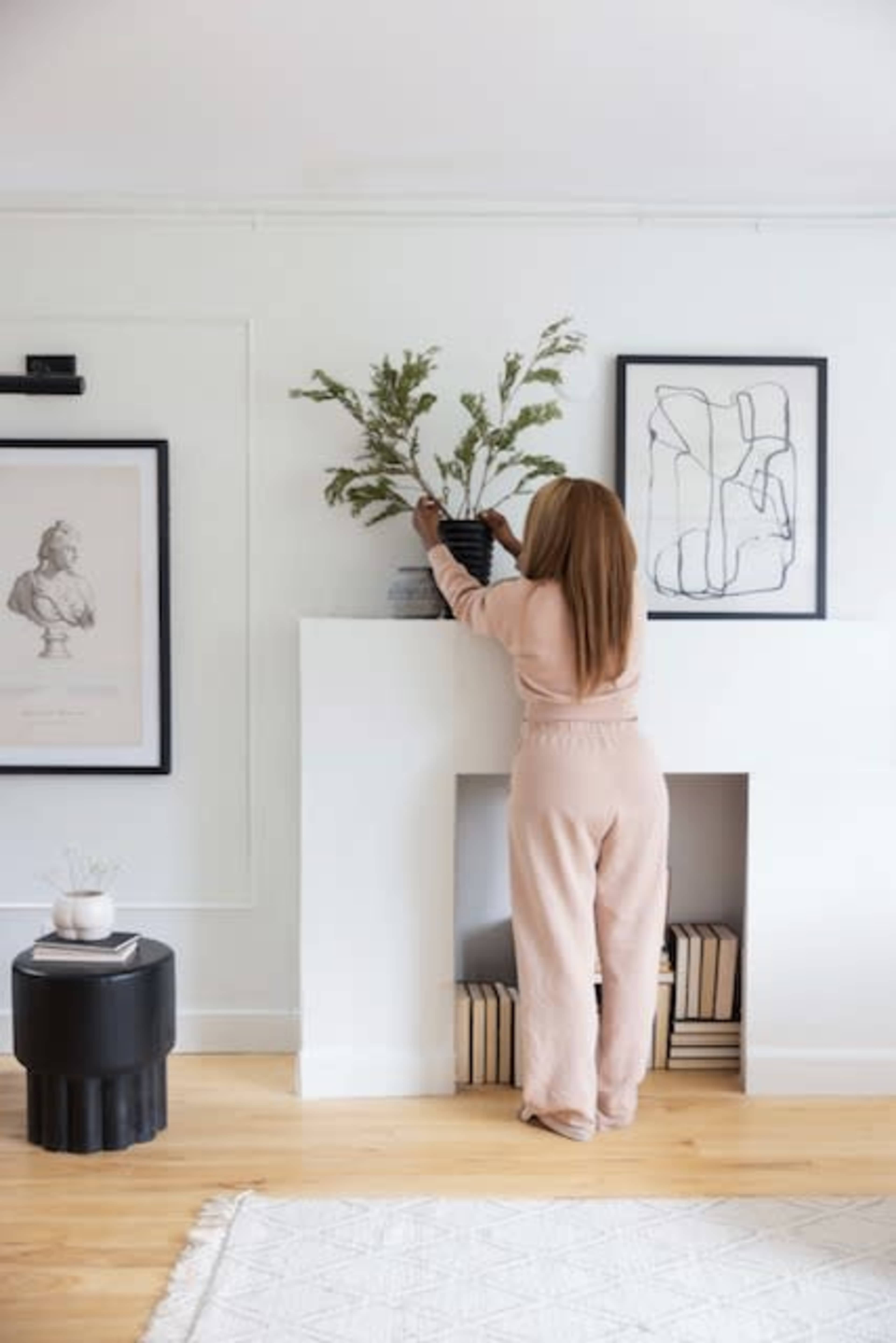 A woman dressed in a pink outfit arranges a plant in a black pot on a white mantel in a modern interior.